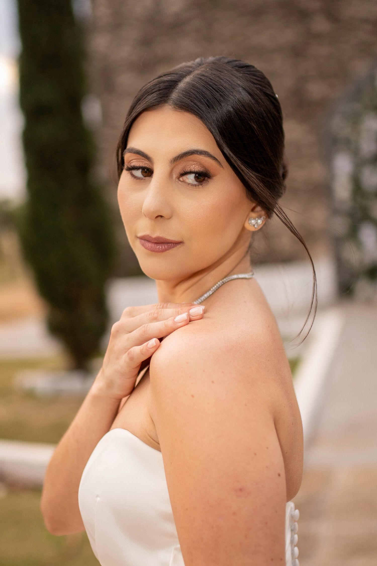 Elegant bride in white strapless gown with pearl earrings and necklace, posing outdoors with hand on shoulder.
