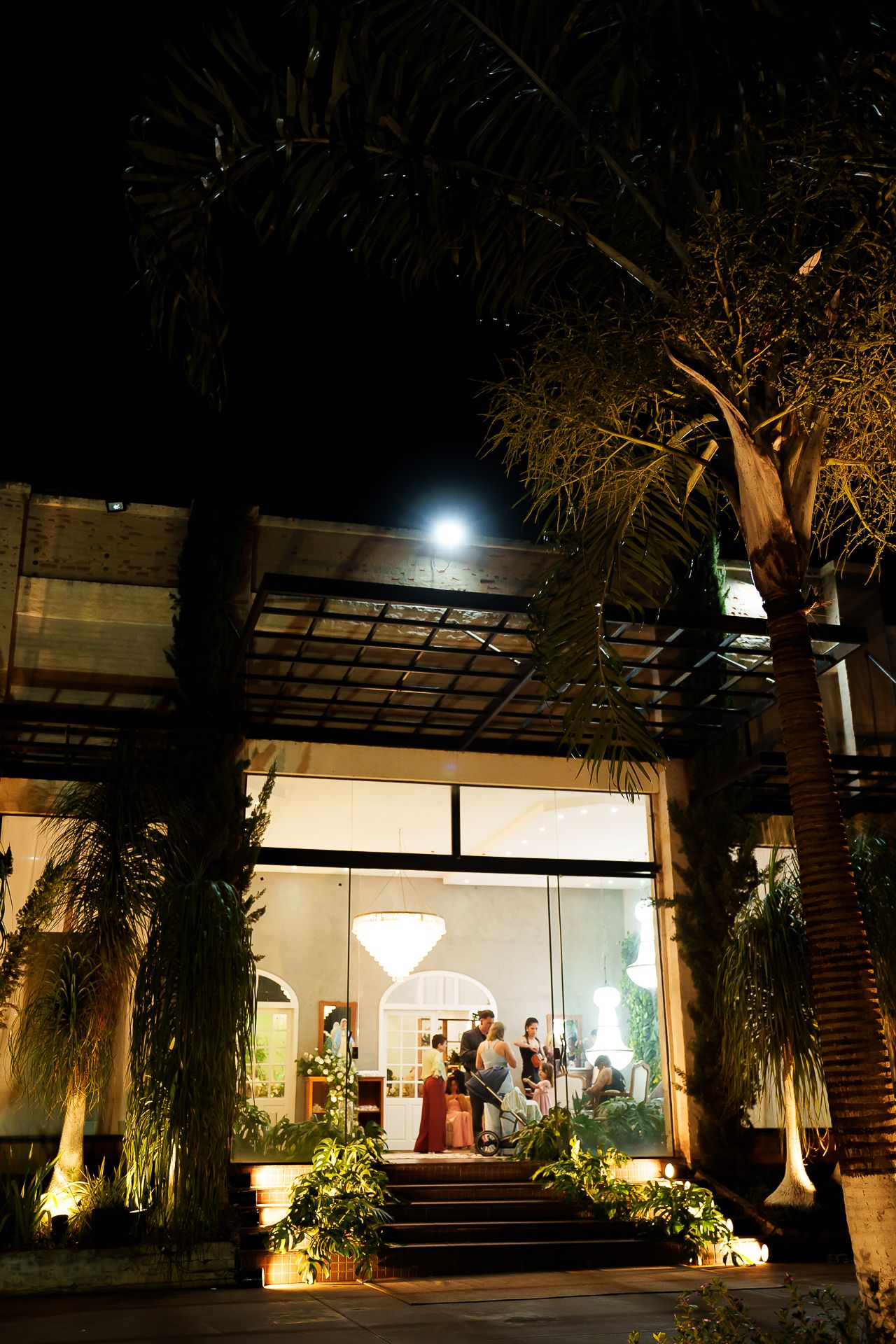 Elegant venue entrance lit at night with lush tropical plants, steps, and guests visible through large glass windows.
