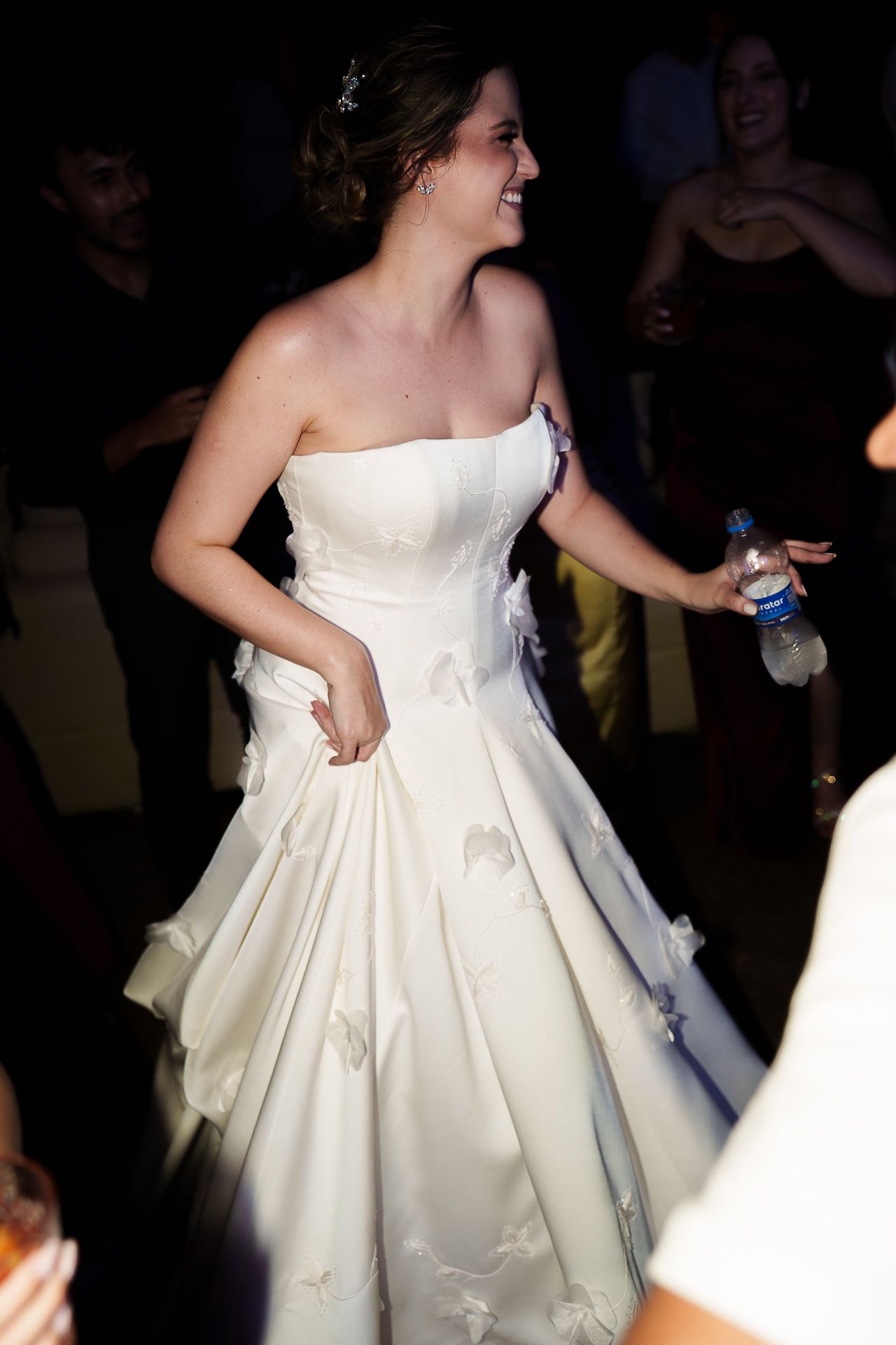 Bride in strapless white ball gown dancing joyfully at wedding reception holding water bottle.