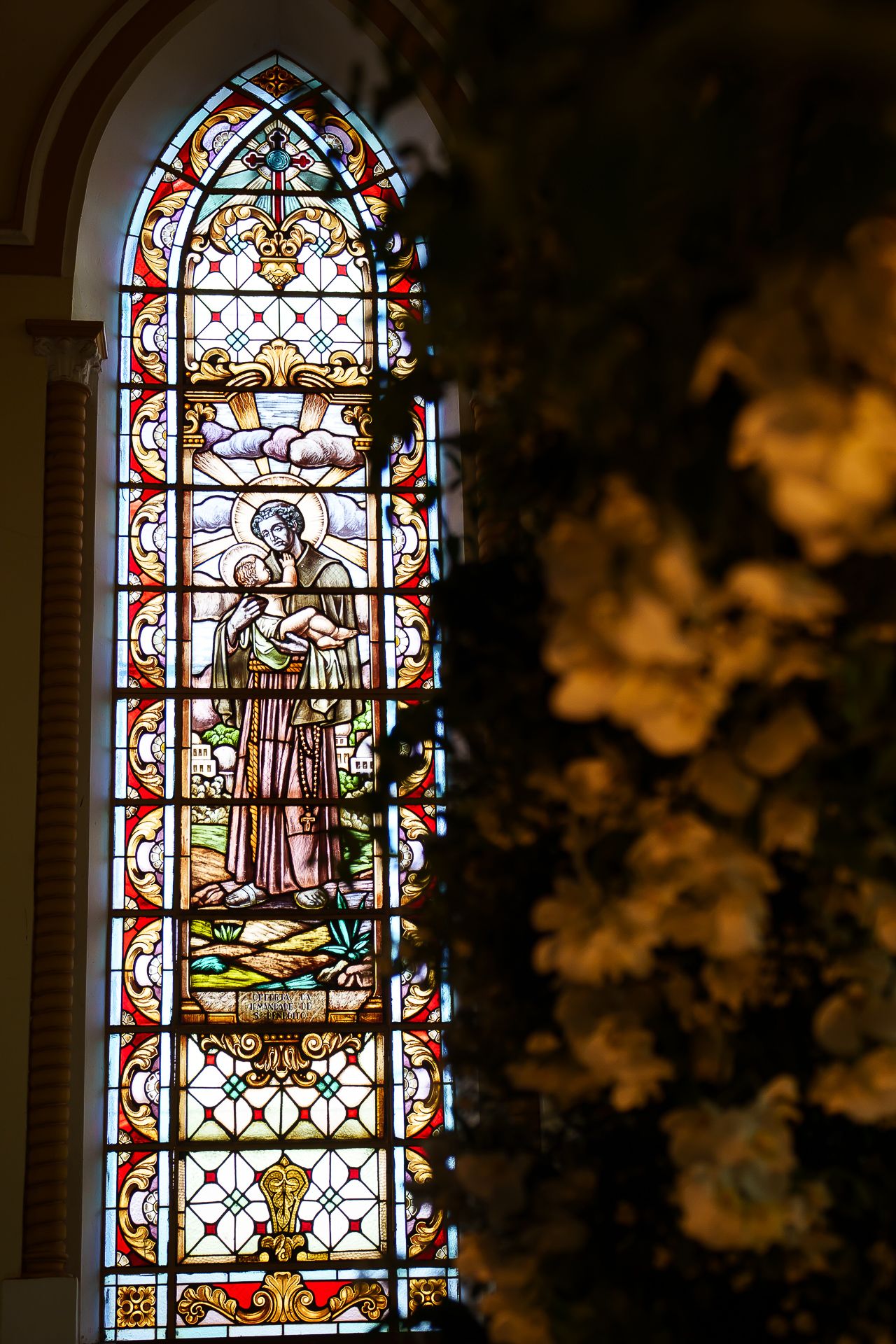 Ornate stained glass church window depicting a religious figure in robes, framed by gothic arches and colorful decorative panels.