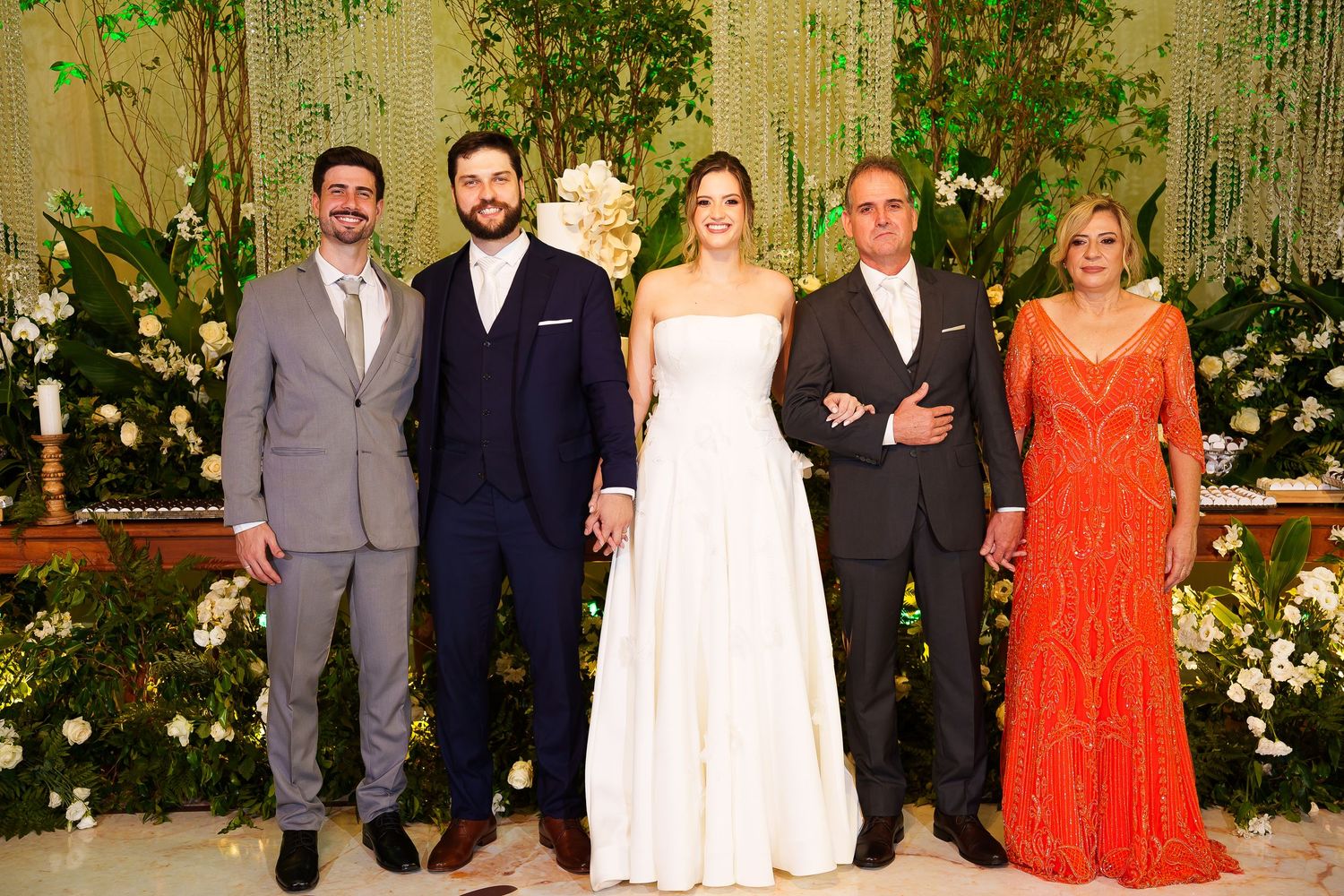 Wedding party of five posing elegantly in front of lush green floral backdrop, bride in white gown flanked by family.