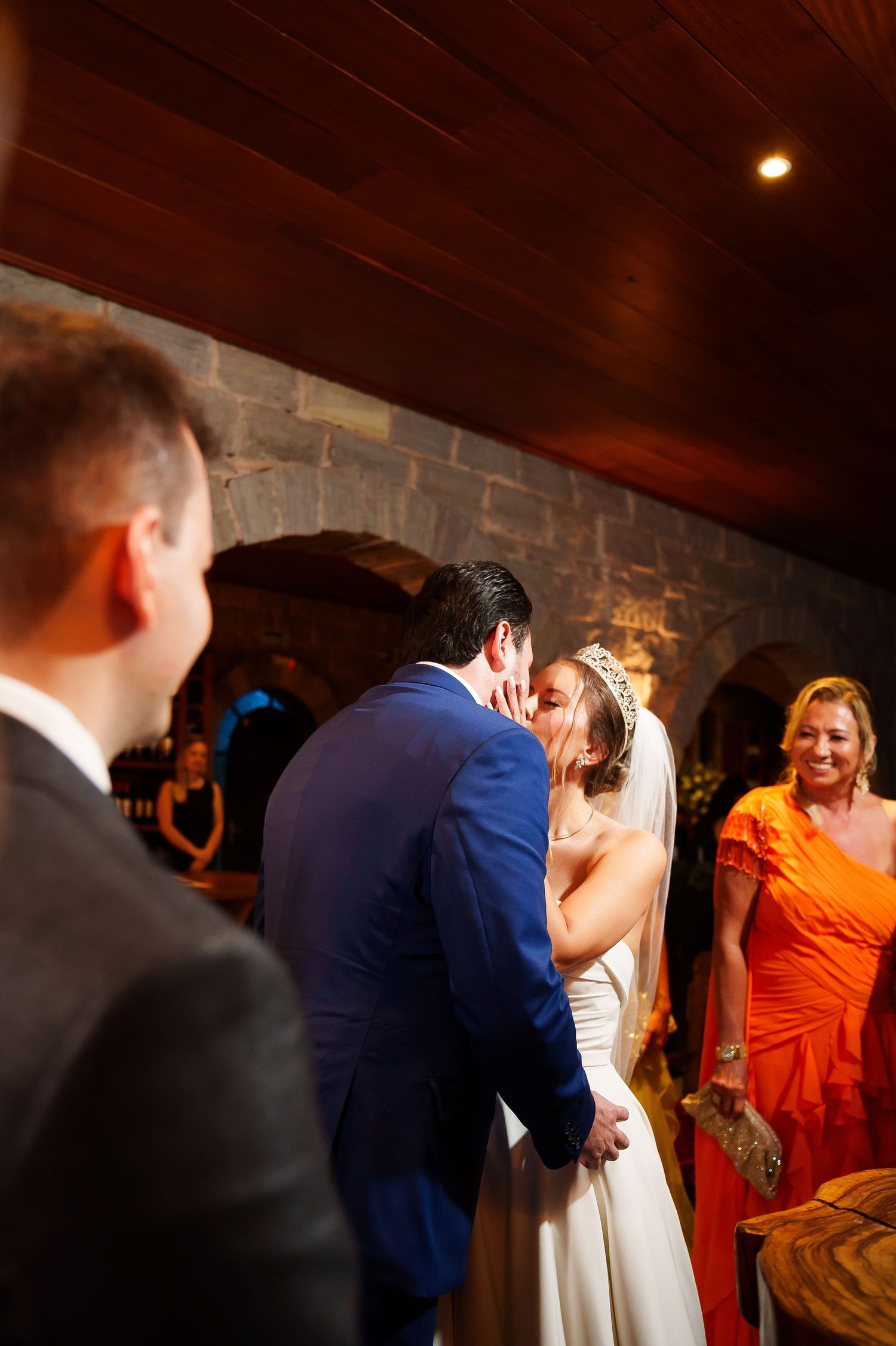 Bride and groom share first kiss at wedding ceremony in stone venue, with guests in orange dress smiling nearby.
