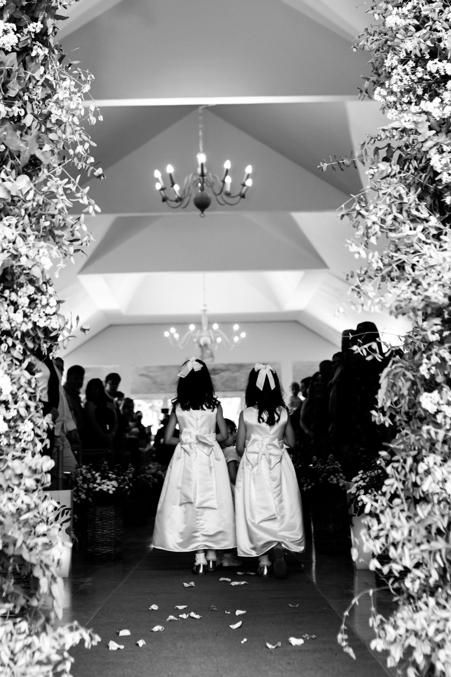 Two flower girls in white dresses walk down a floral-lined aisle at an elegant wedding ceremony.
