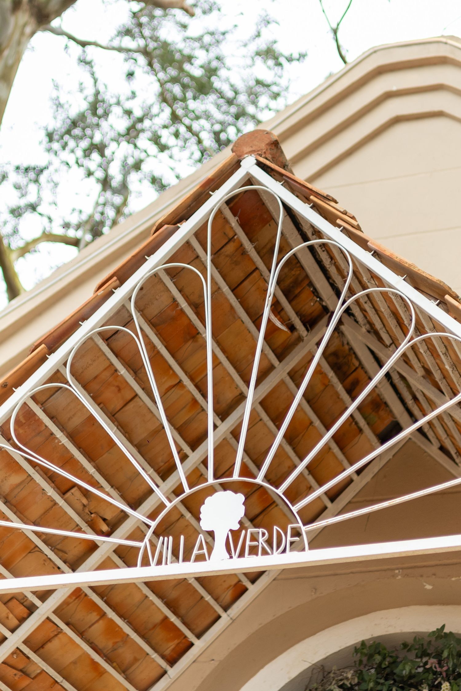 White ornate iron gate with Villa Verde sign on a terracotta-roofed entrance surrounded by lush trees.