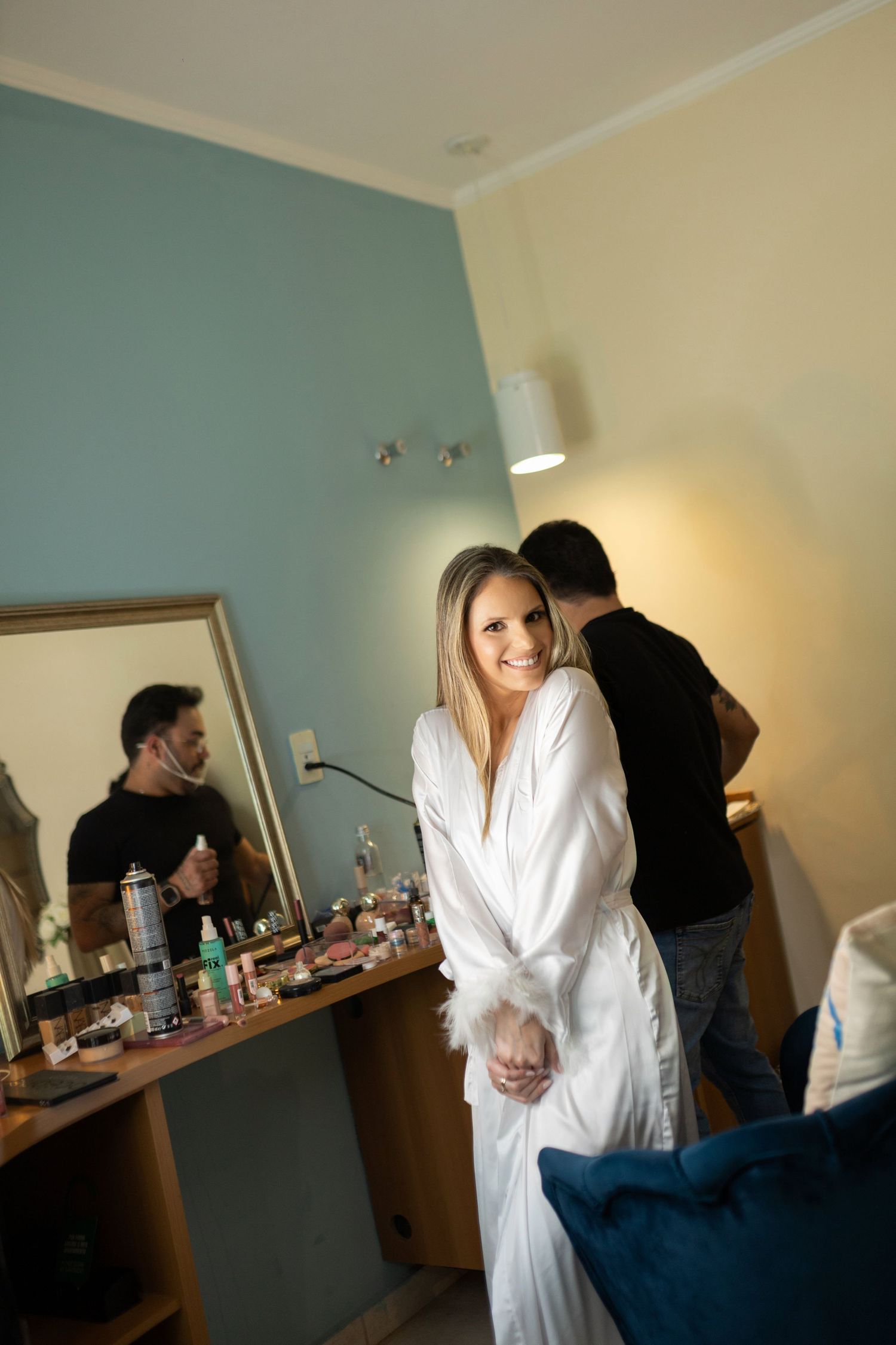 Smiling bride in white robe poses in hotel room while makeup artist prepares at vanity mirror.
