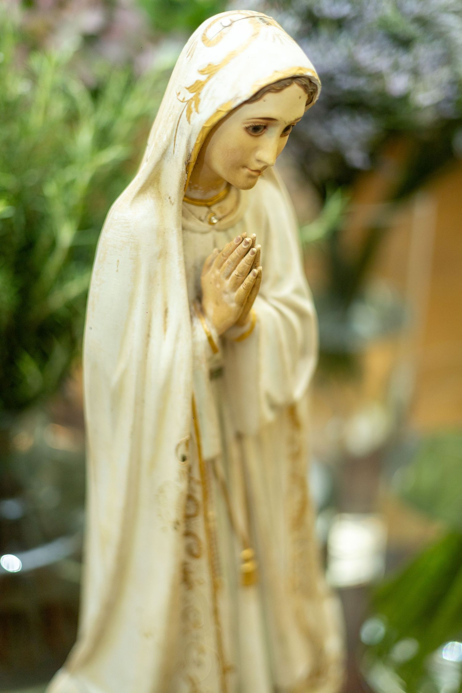 White and gold Virgin Mary statue with hands in prayer position, surrounded by green foliage background.