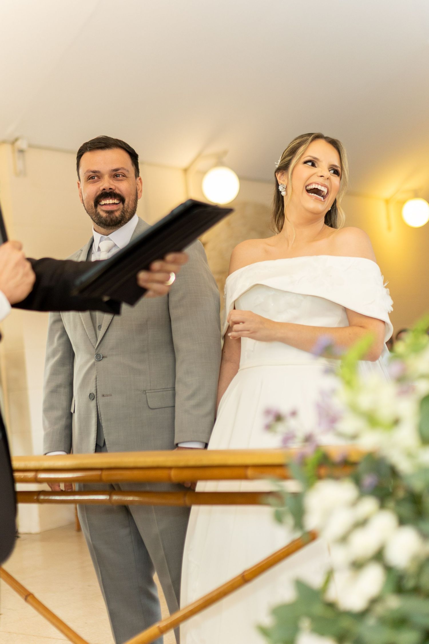 Smiling bride in off-shoulder white gown and groom in gray suit laughing joyfully during wedding ceremony indoors.