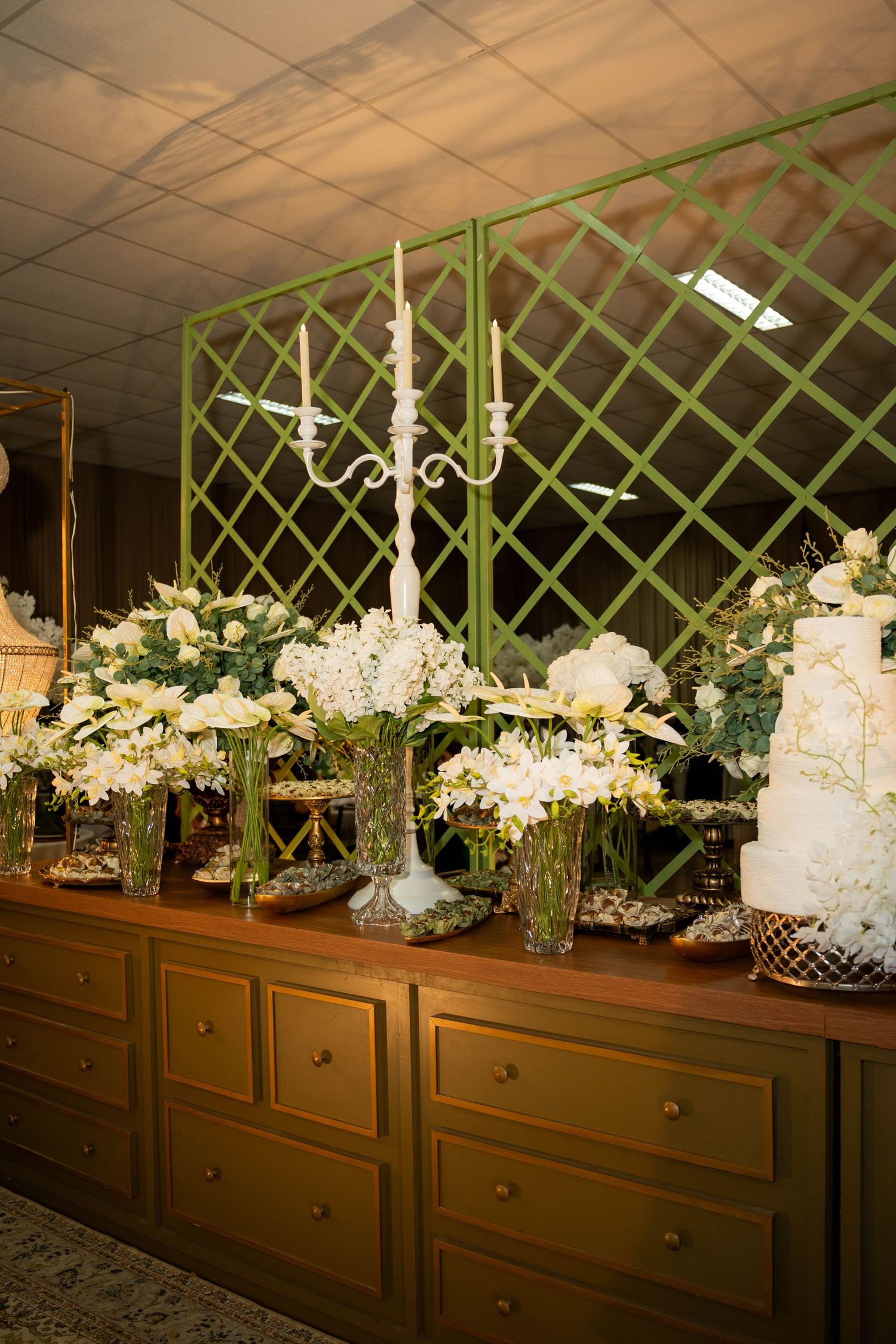 Wedding dessert table with white floral arrangements, candelabra centerpiece, and tiered cake against green lattice backdrop.