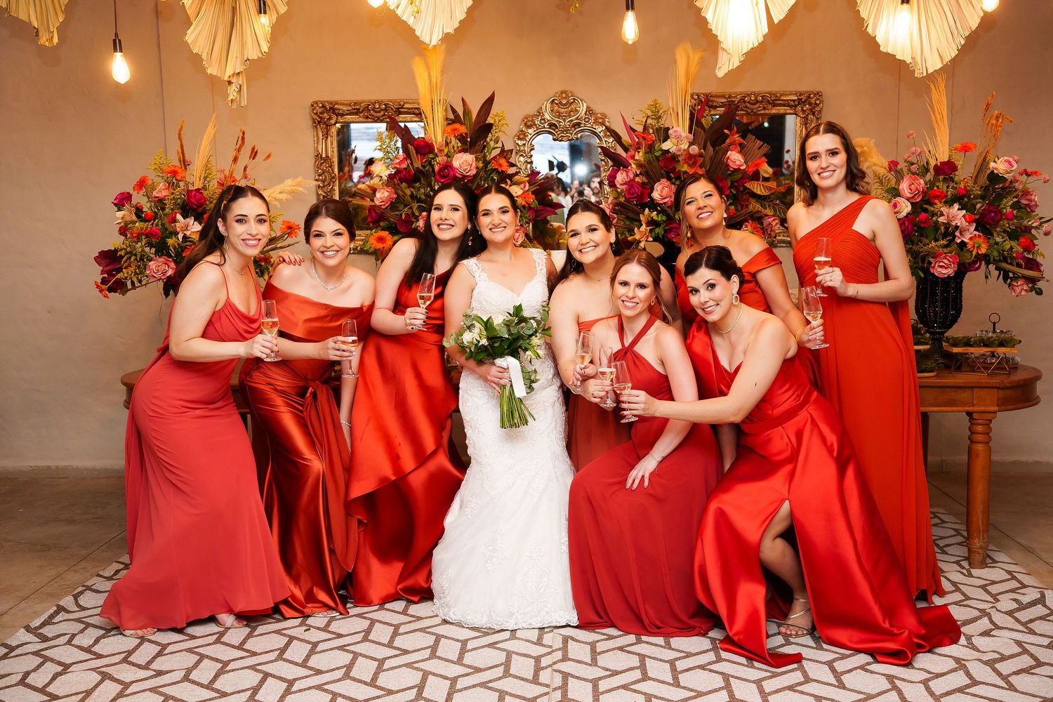Bride in white gown poses with bridesmaids in red satin dresses, holding bouquets amid floral arrangements.