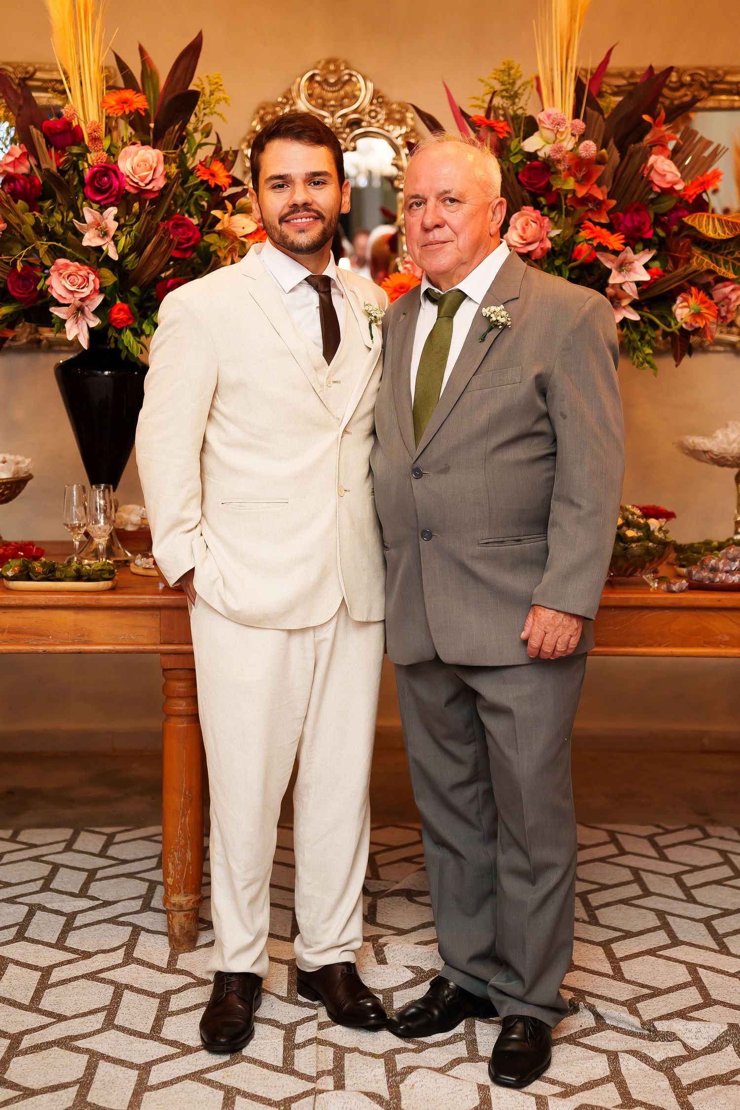 Two men in suits pose together at a wedding, surrounded by colorful floral arrangements.