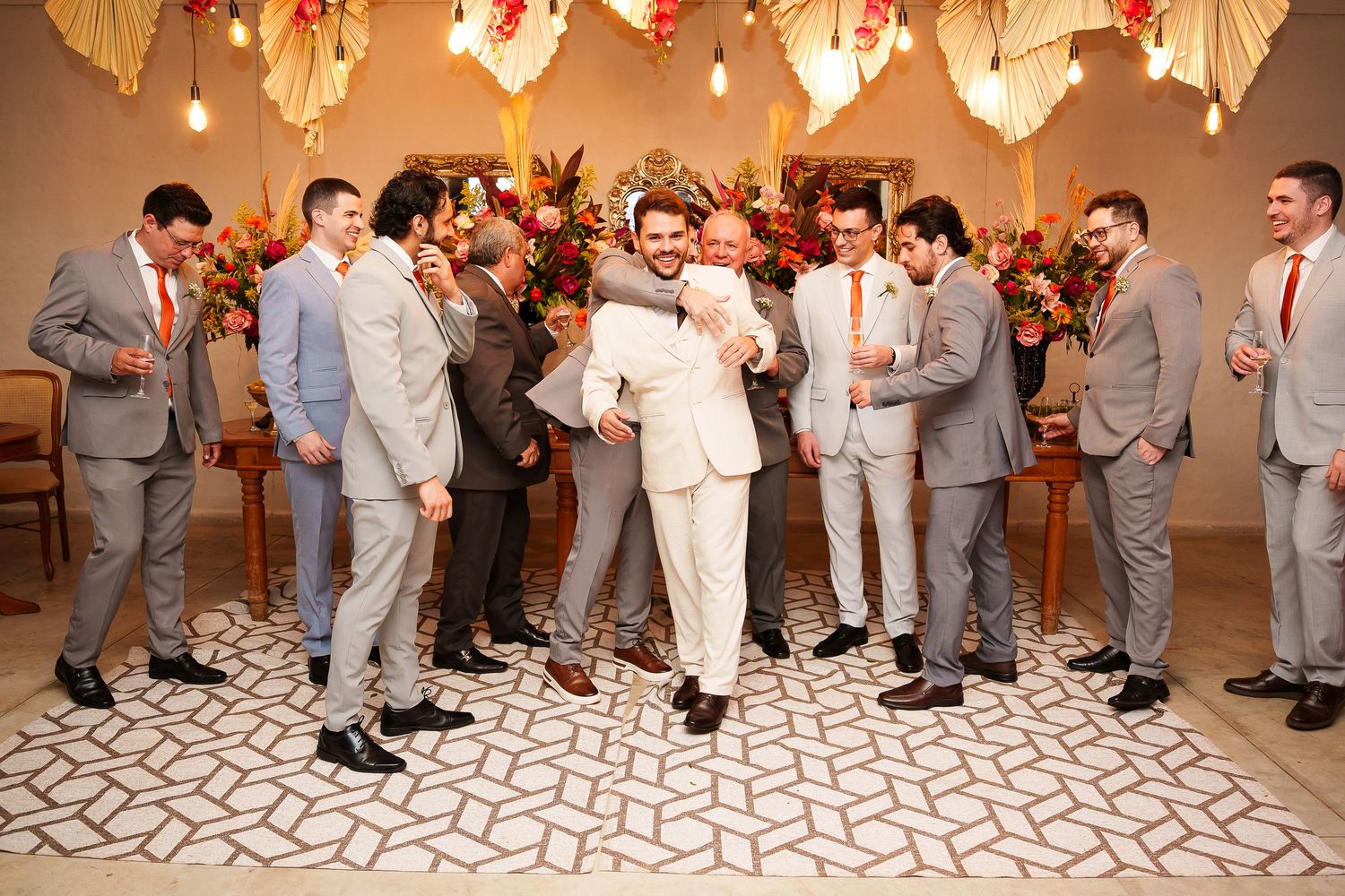 Groom in white suit celebrates with groomsmen in gray suits at elegant wedding ceremony venue with floral decor.