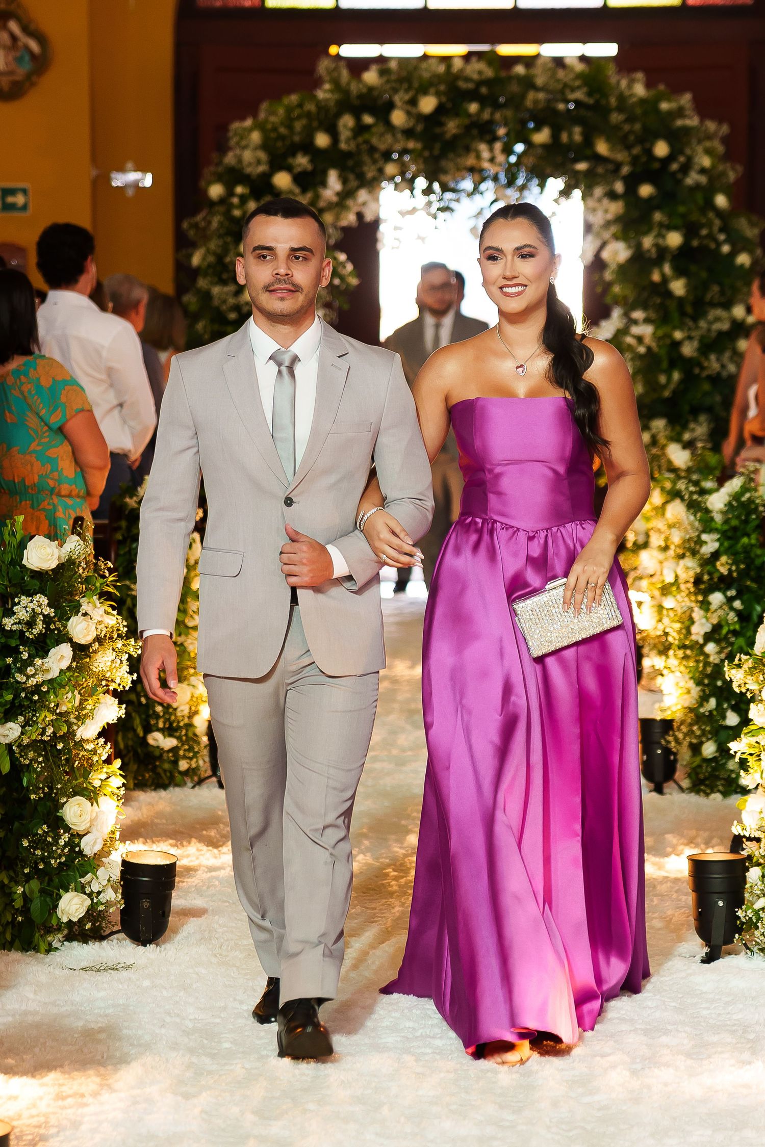 Elegant couple walking down wedding aisle, man in gray suit, woman in purple strapless gown holding clutch.