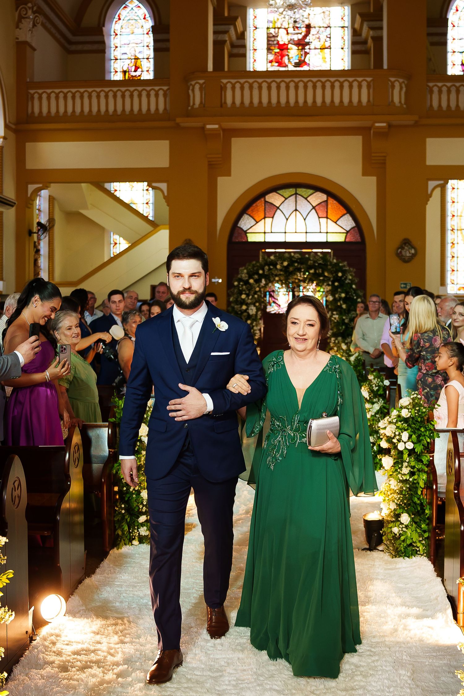 Groom escorts woman in green dress down flower-lined church aisle surrounded by wedding guests.