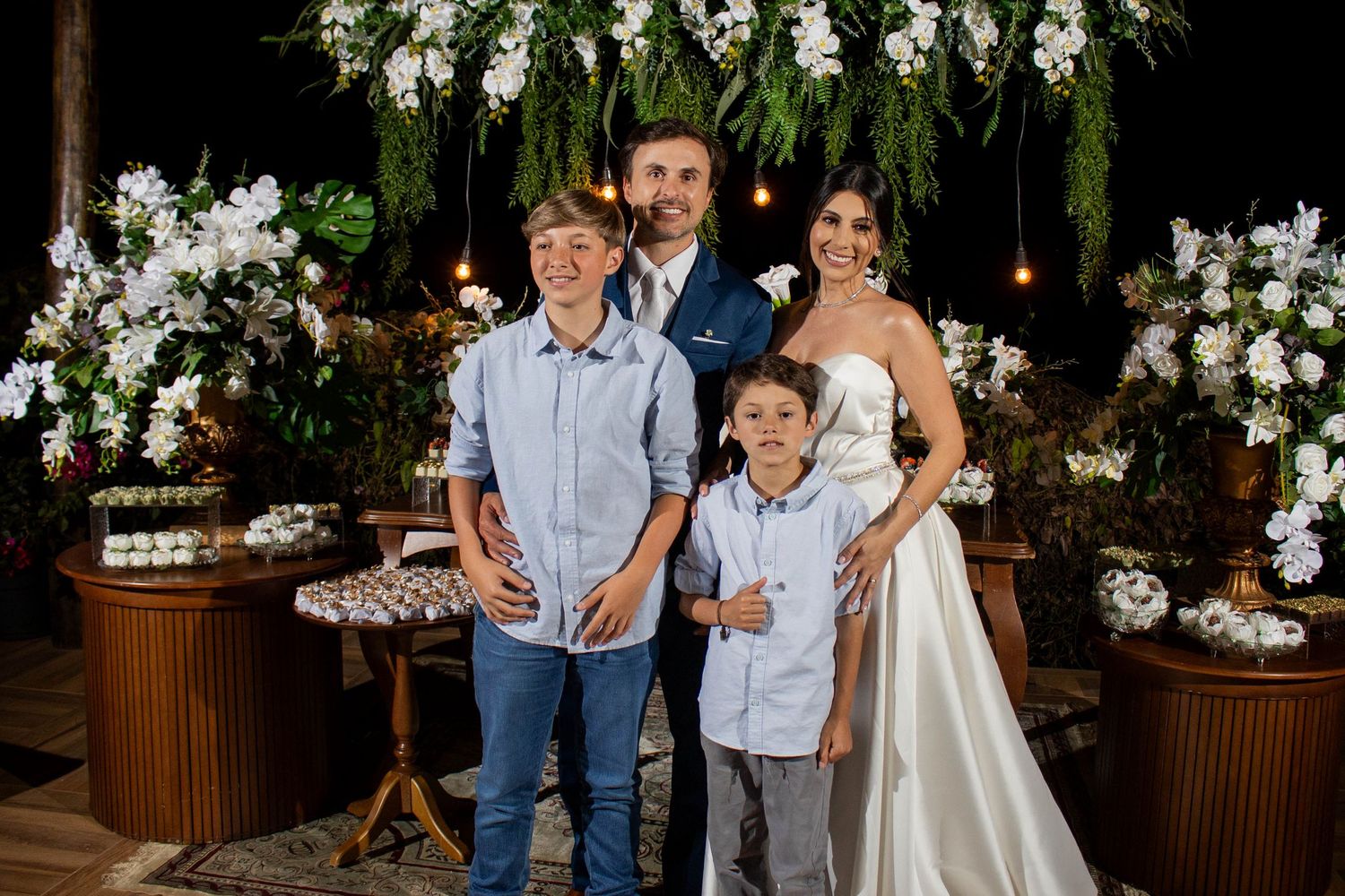 Family of four poses together at an elegant outdoor wedding reception with white floral decorations and dessert table.