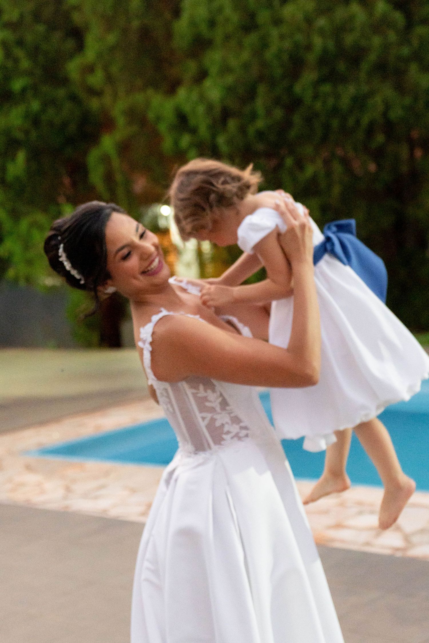 Smiling bride in white gown lifts young flower girl in white dress near a pool outdoors.