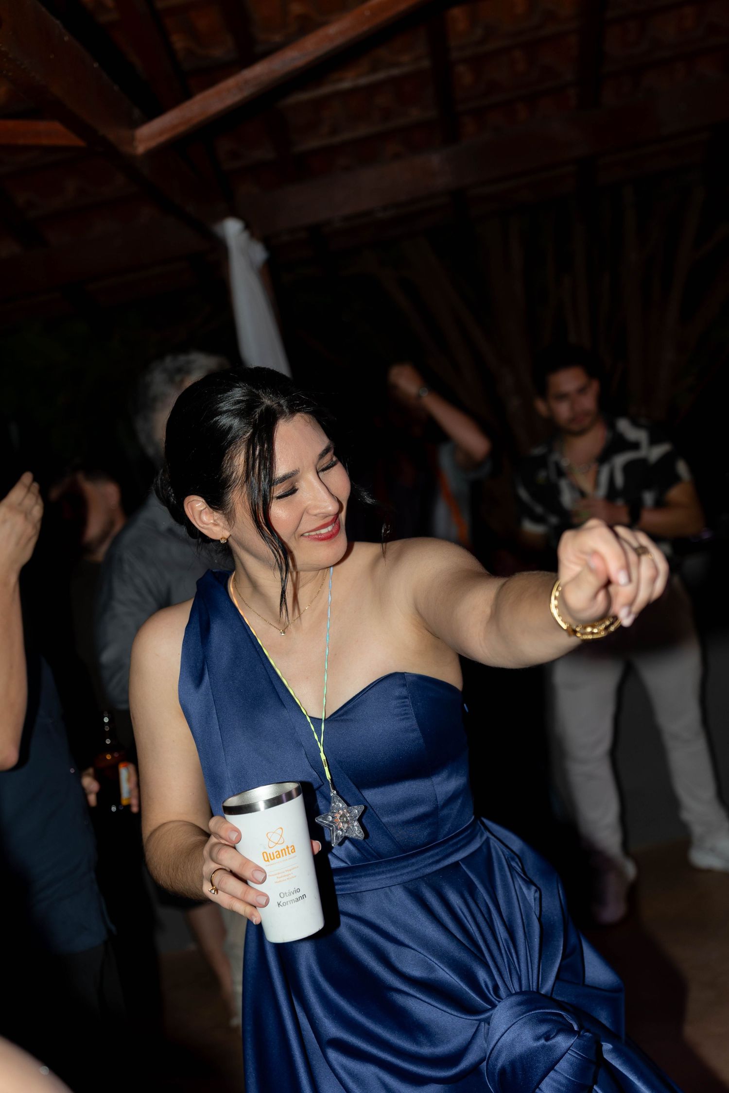 Woman in blue satin gown dancing joyfully at a party, holding a tumbler cup while reaching out her hand.
