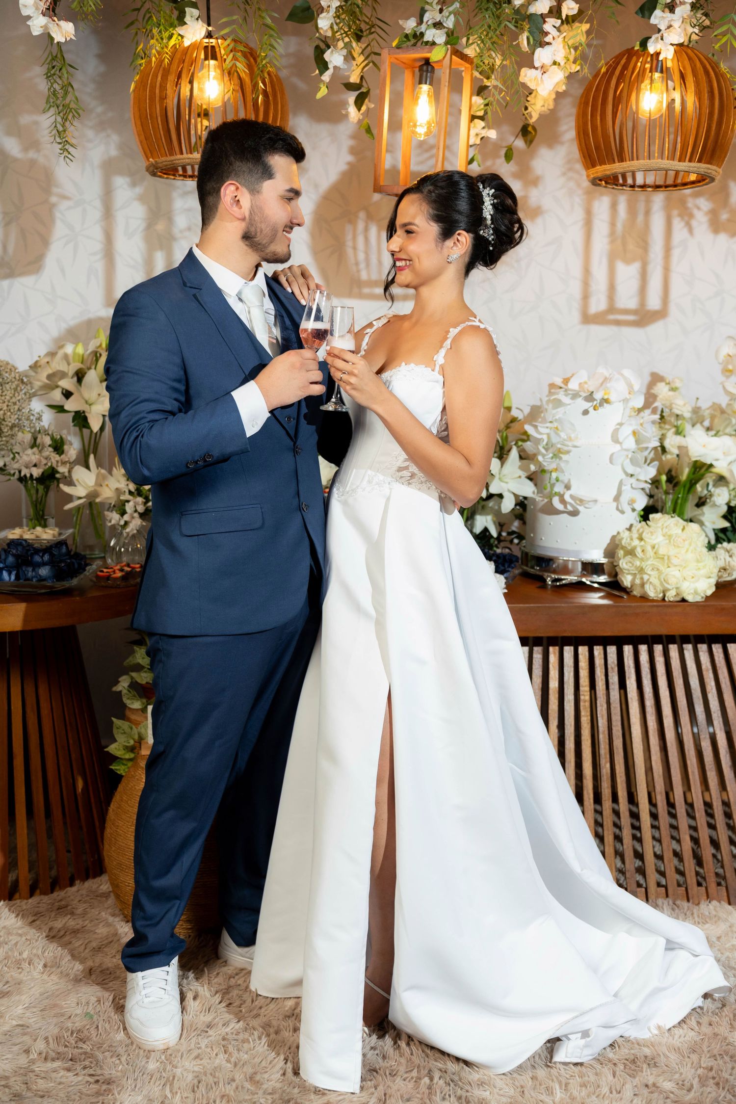 A bride and groom share a champagne toast at their elegant wedding reception surrounded by white floral arrangements.