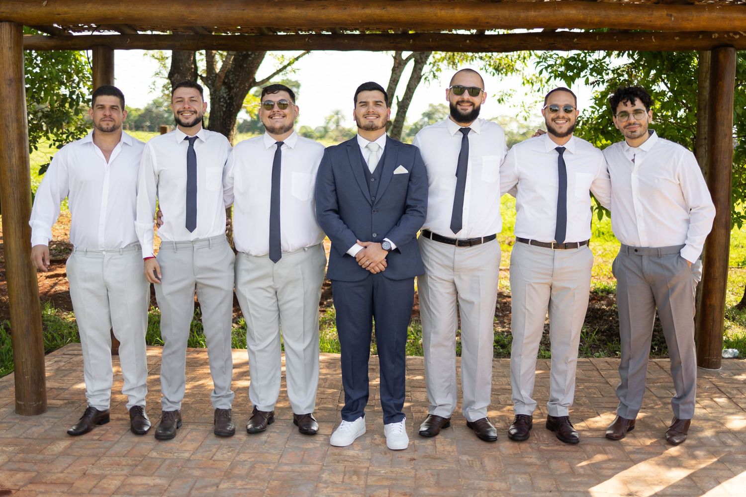 Groom in navy suit poses with six groomsmen in white shirts and ties outdoors at a wedding venue.