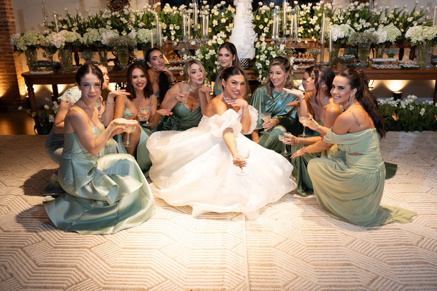 Bride in white gown sits on dance floor surrounded by bridesmaids in sage green dresses at elegant floral wedding reception.