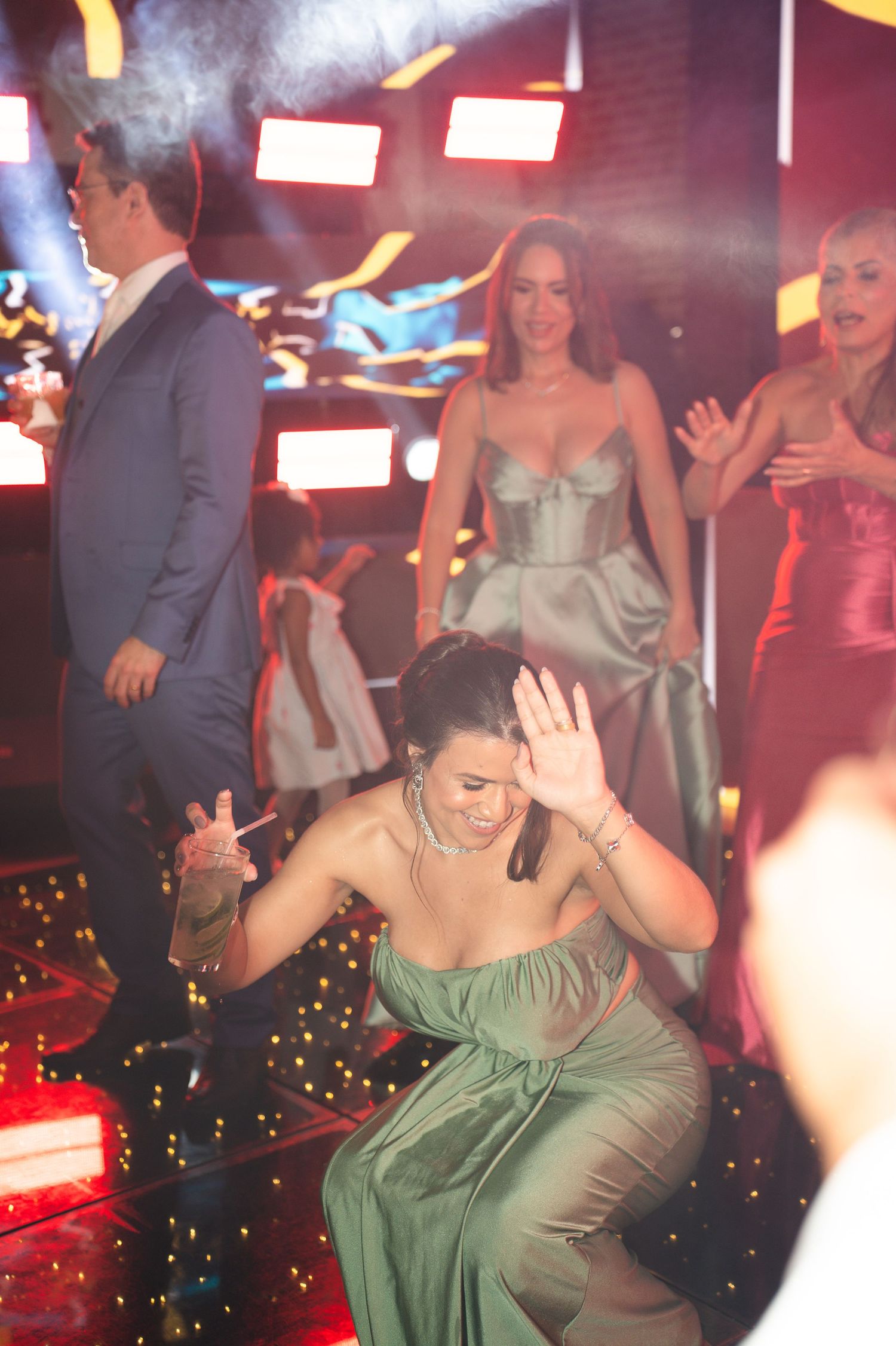 Woman in green dress dancing energetically on a lit dance floor surrounded by elegantly dressed guests at a party.