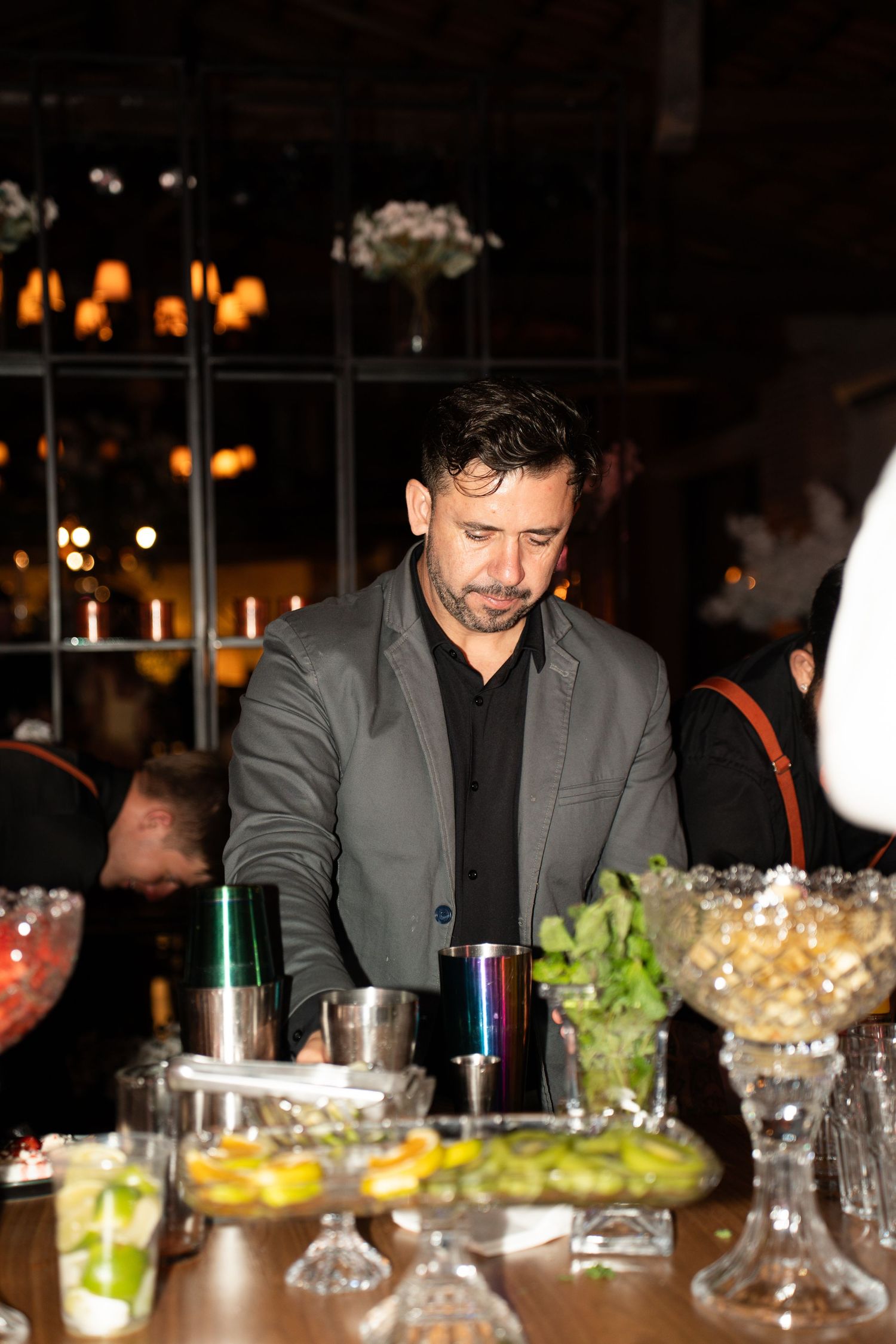Bartender in gray blazer preparing cocktails at a bar with fresh fruit garnishes and glassware in dimly lit venue.