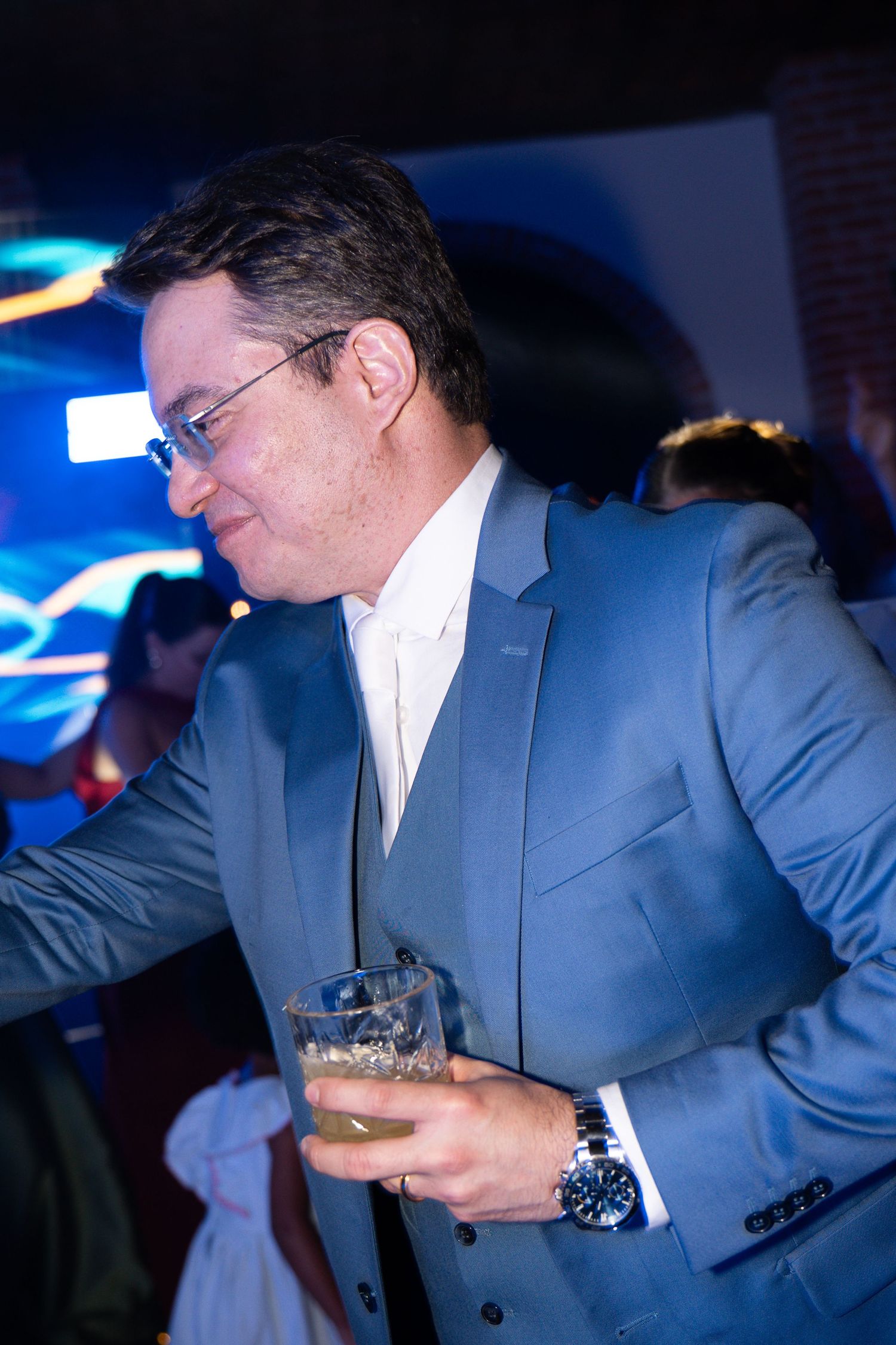 Man in blue suit holding whiskey glass at evening event with blue ambient lighting.
