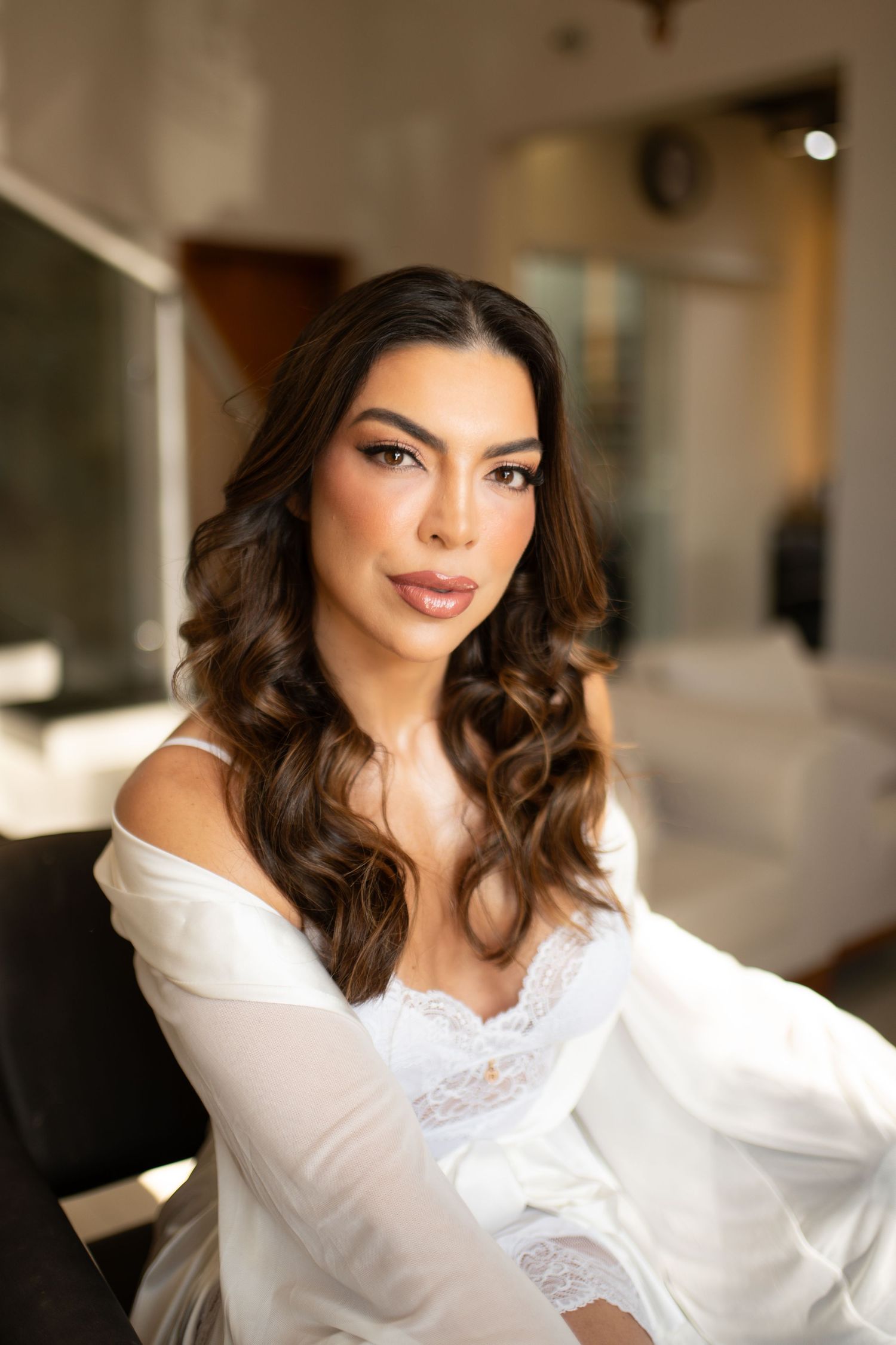 A woman with long curly brunette hair wearing a white off-shoulder lace robe sits elegantly in a salon chair.
