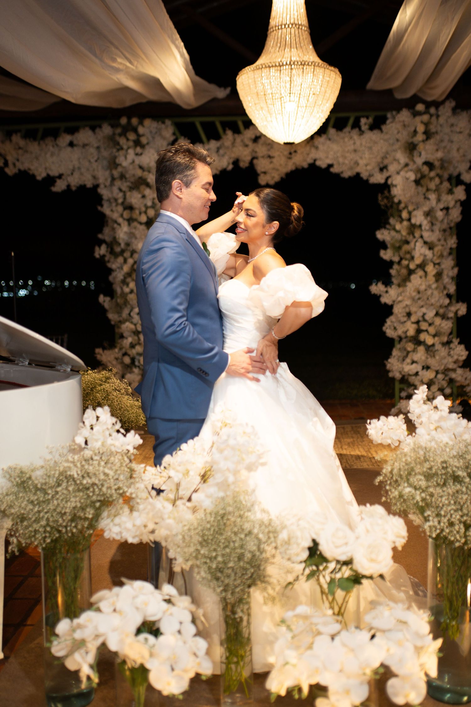 Bride and groom share a tender moment under a chandelier surrounded by white floral arrangements at an elegant outdoor night wedding.