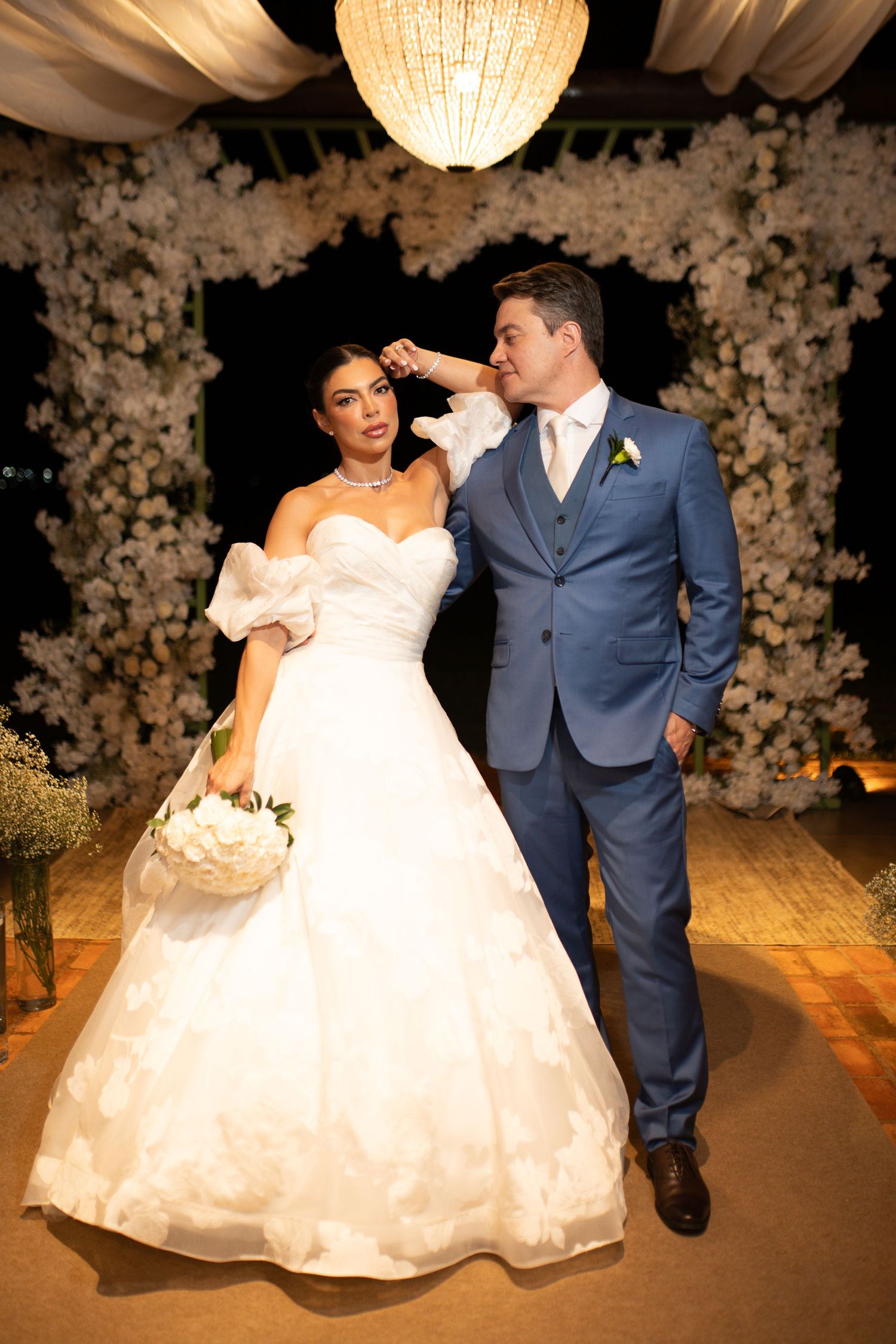 Bride in white ball gown and groom in blue suit pose together under floral arch at elegant evening wedding ceremony.