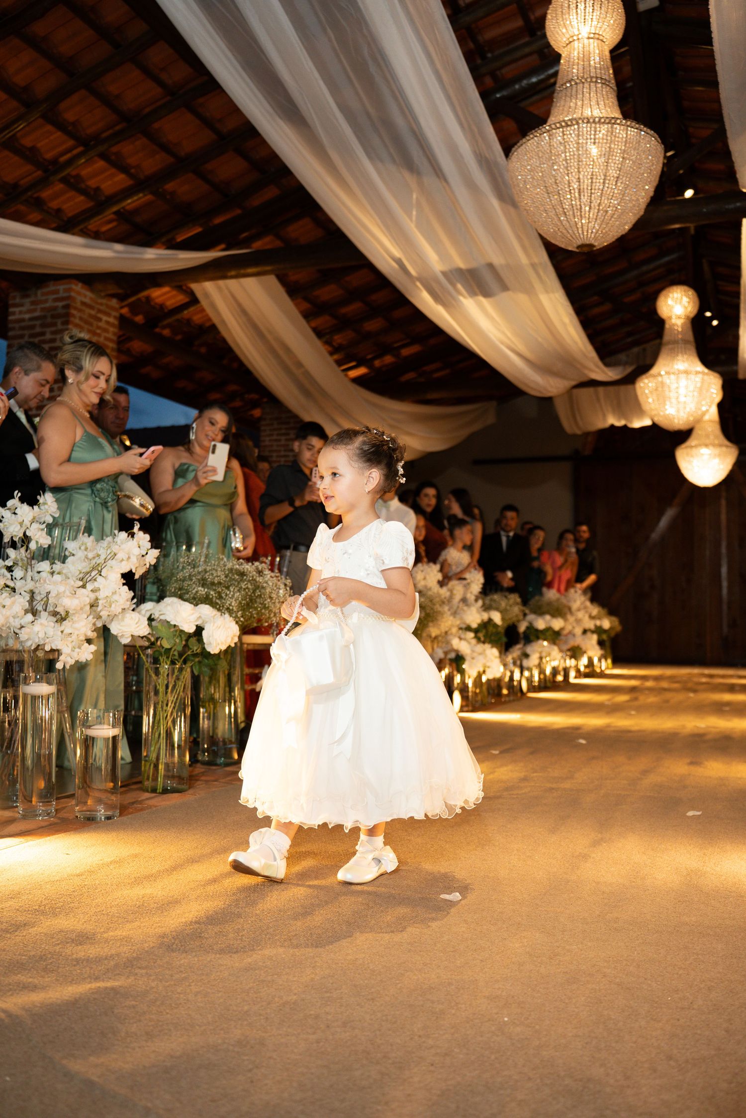 A flower girl in a white dress walks down the aisle at an elegant wedding reception with chandeliers and floral arrangements.