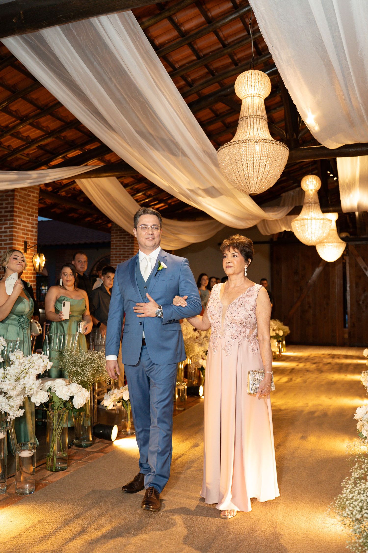Groom in blue suit walks down wedding aisle with elegantly dressed older woman amid chandeliers and white draping.