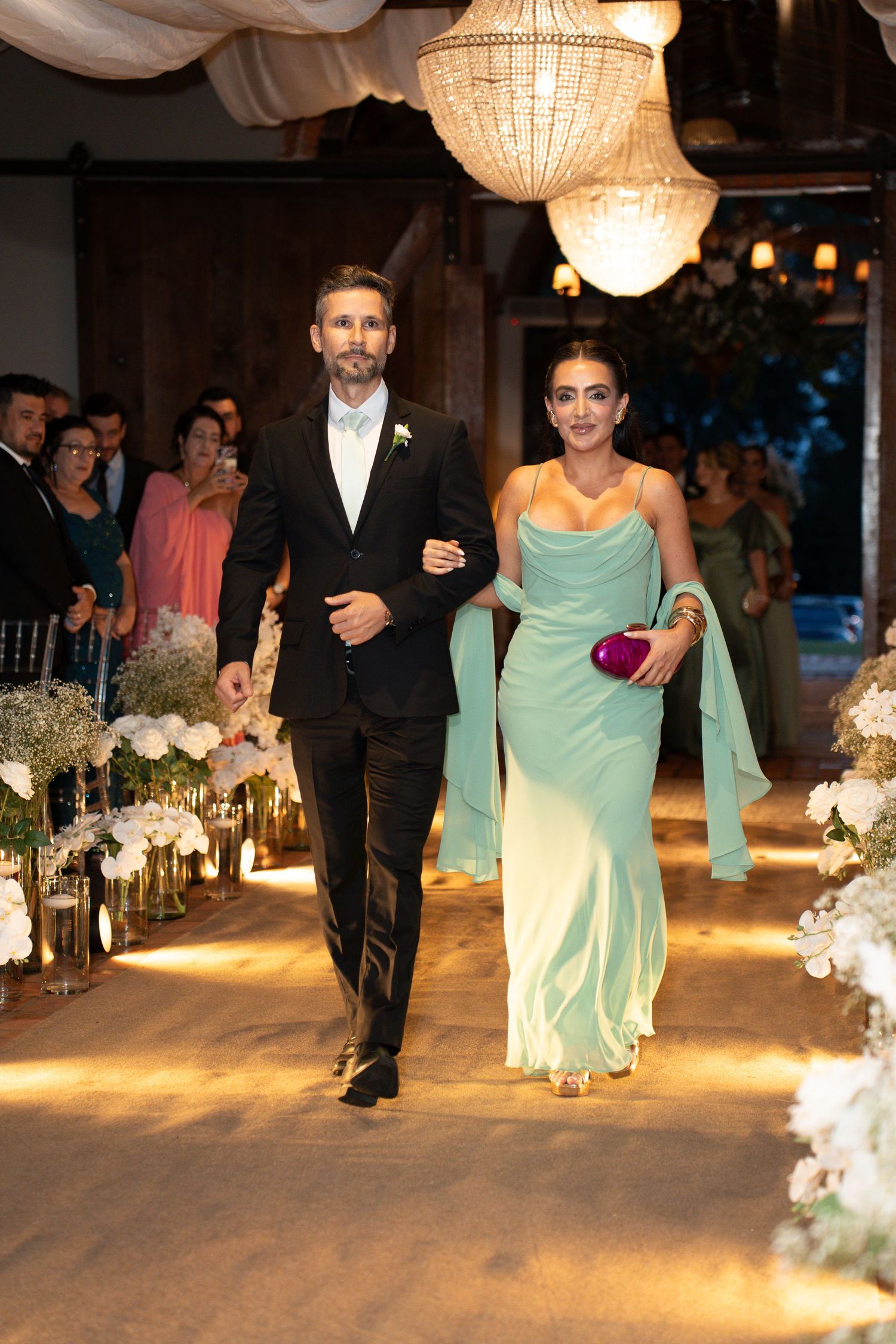 Elegant couple walking down wedding aisle, man in black suit, woman in mint green gown, crystal chandeliers above.