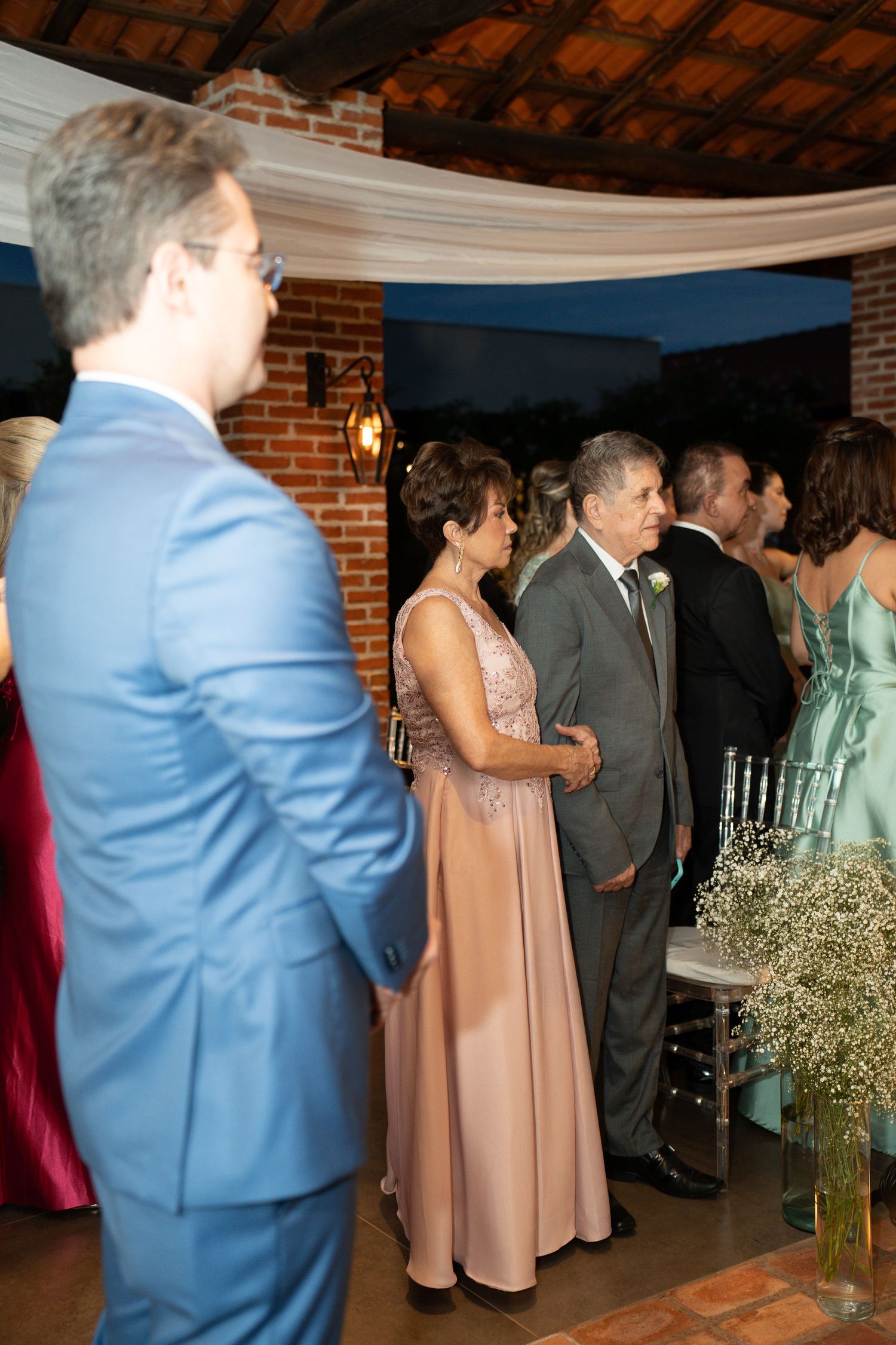 Elegant wedding guests in formal attire, including a blue suit and pink gown, gathered at an indoor venue with brick walls.