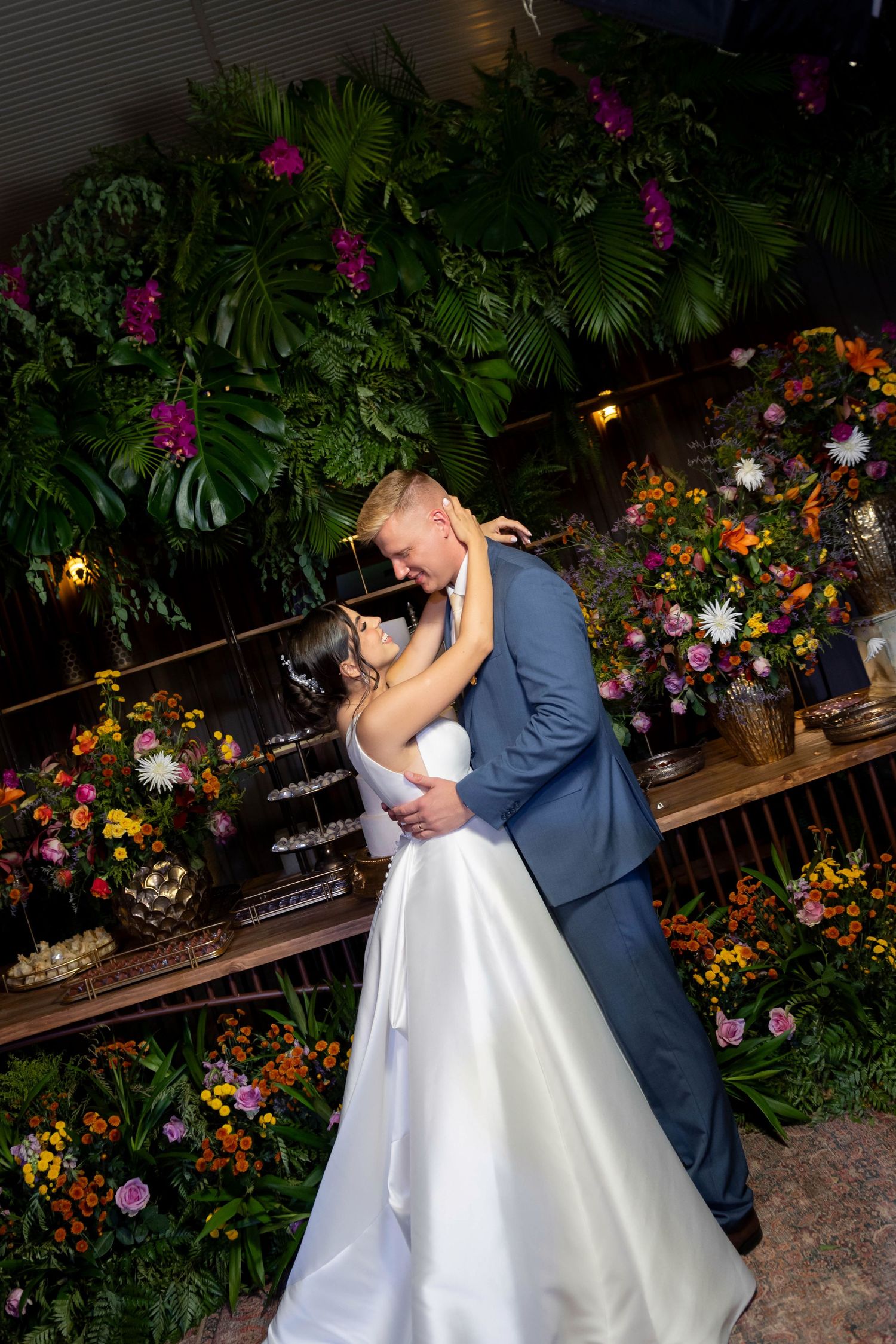 Bride and groom share a romantic first dance embrace surrounded by lush tropical greenery and colorful floral arrangements.