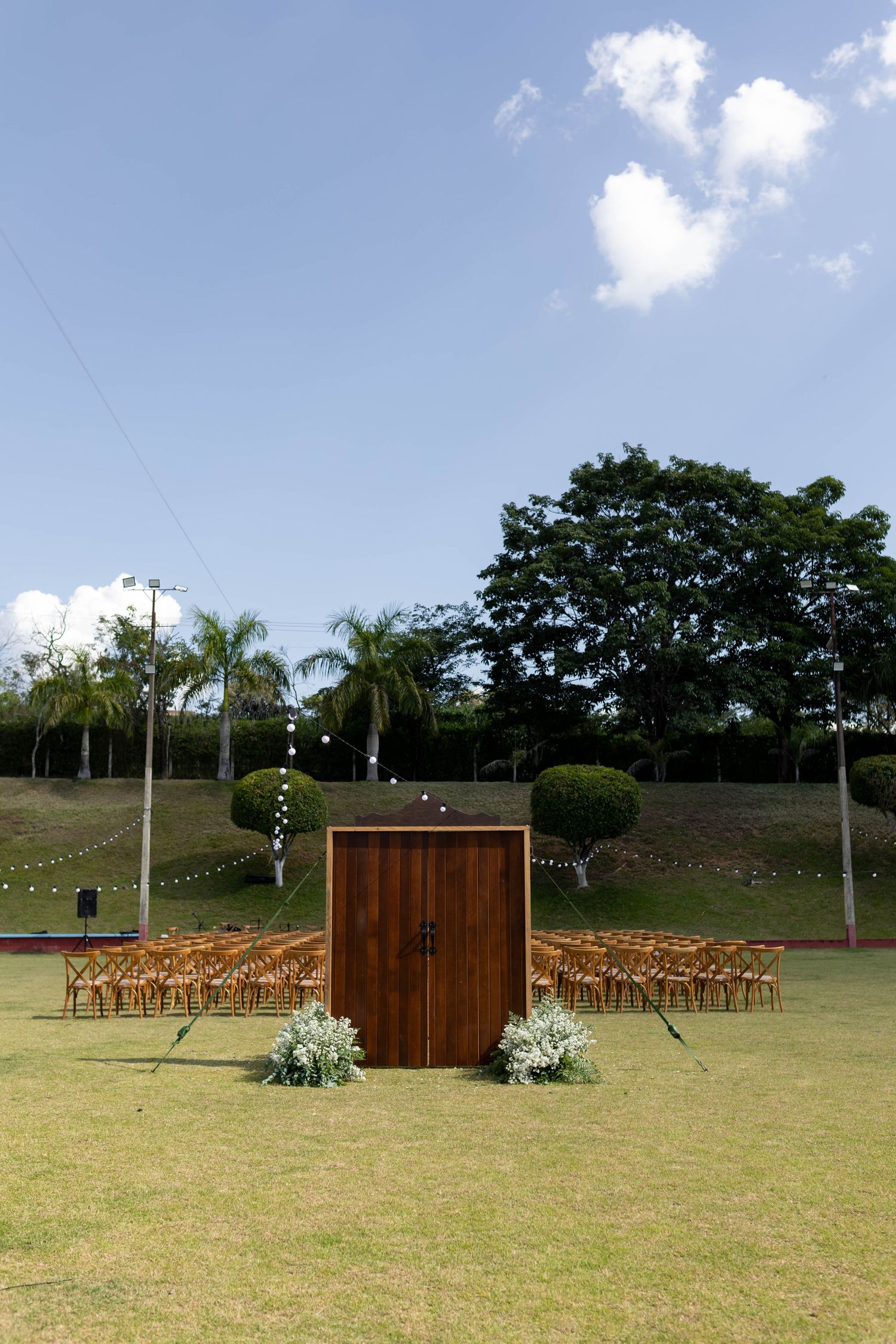Outdoor wedding ceremony setup with wooden doors, floral arrangements, and rows of chairs on green lawn surrounded by palm trees.