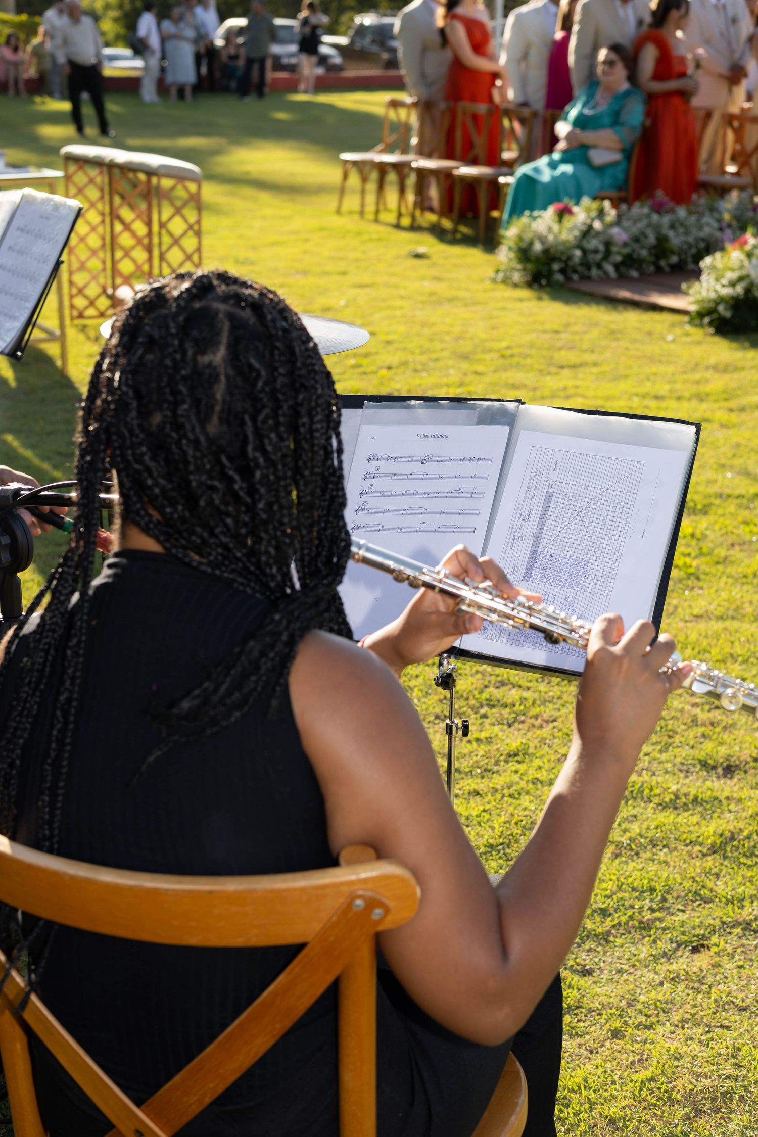 Woman with braids plays flute outdoors at a garden wedding ceremony, reading sheet music on a stand.