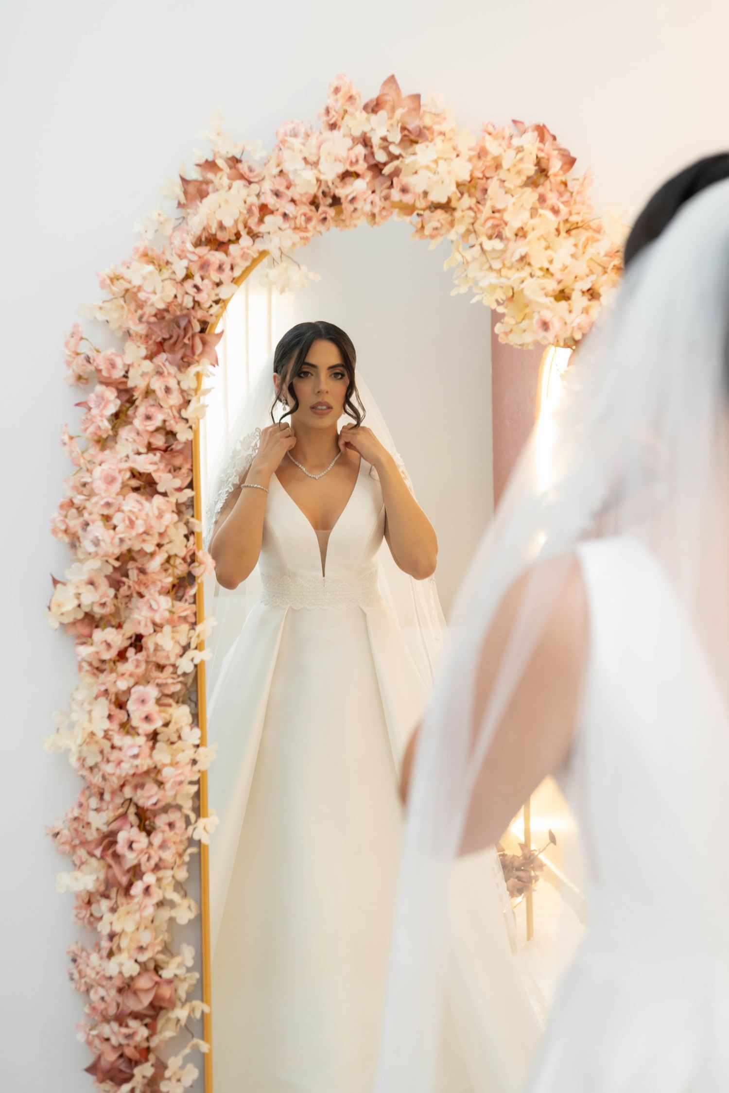 Bride in white gown adjusts necklace before floral arch mirror, reflecting elegance on her wedding day.