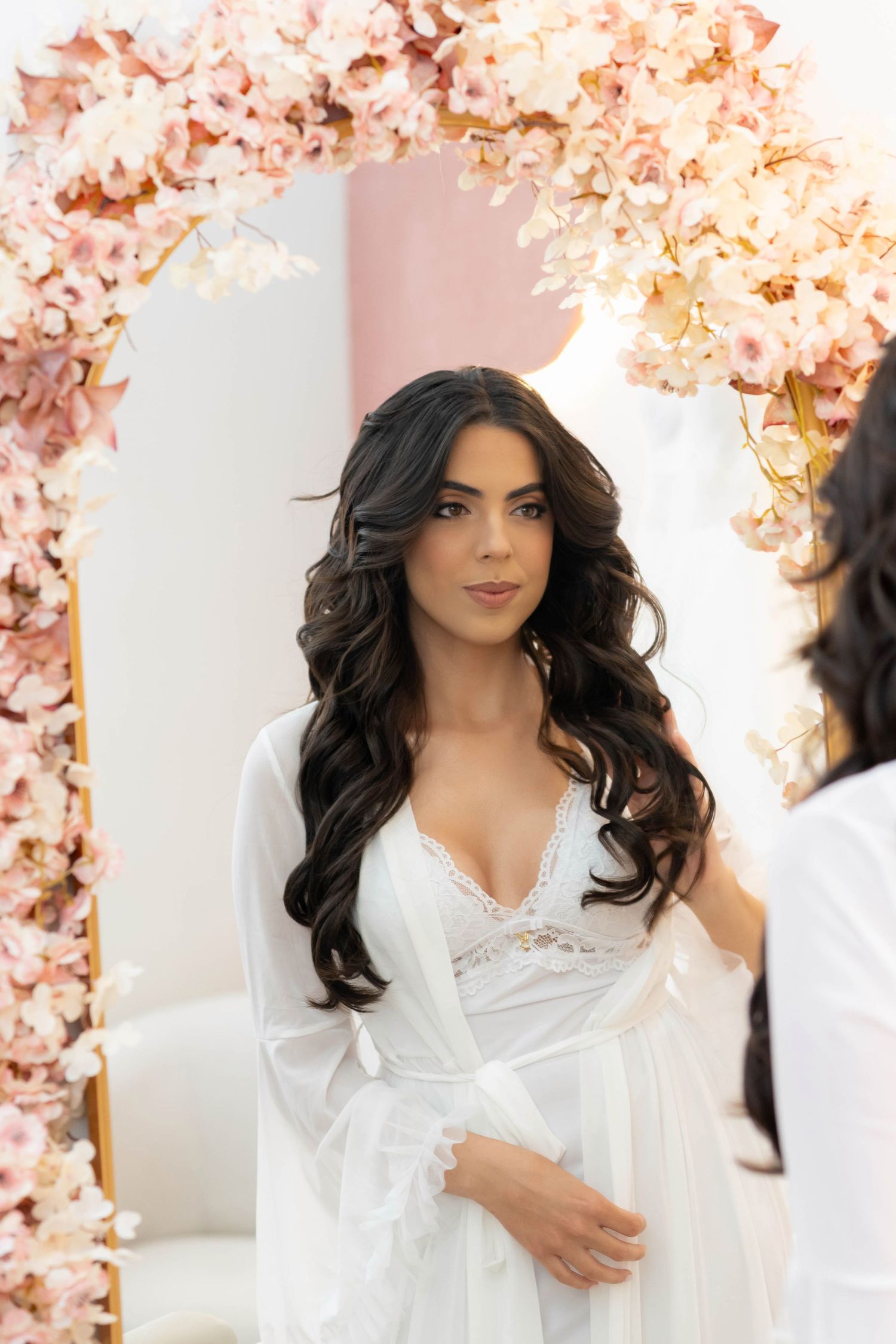 Bride in white lace robe with long dark wavy hair standing before a floral arch mirror.