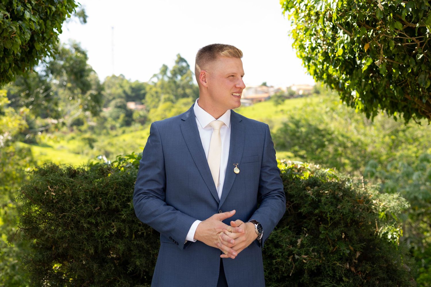 Smiling man in blue suit standing outdoors among green trees and shrubs on a sunny day.