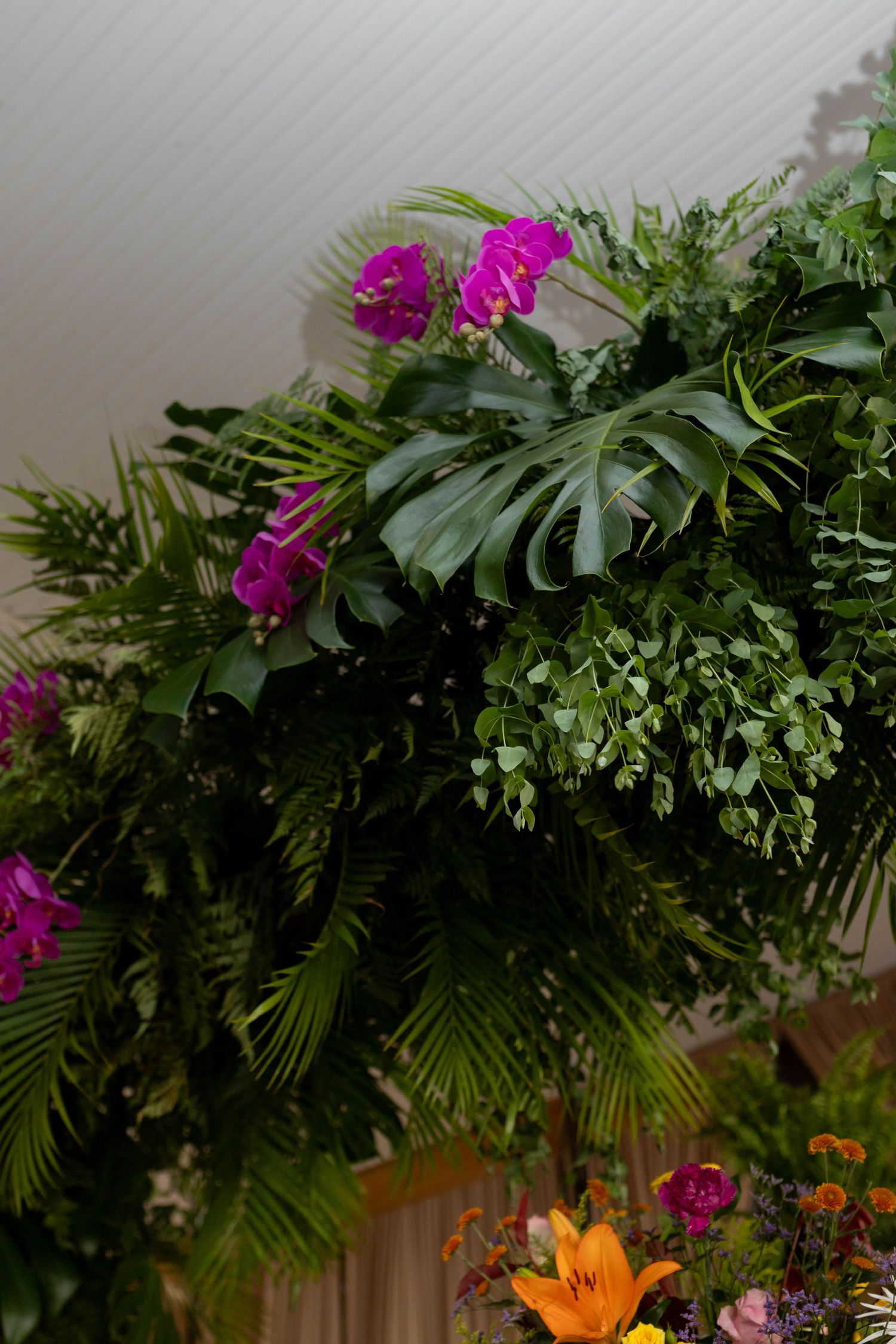 Lush tropical floral arrangement featuring purple orchids, monstera leaves, palm fronds, and orange lilies for an elegant display.