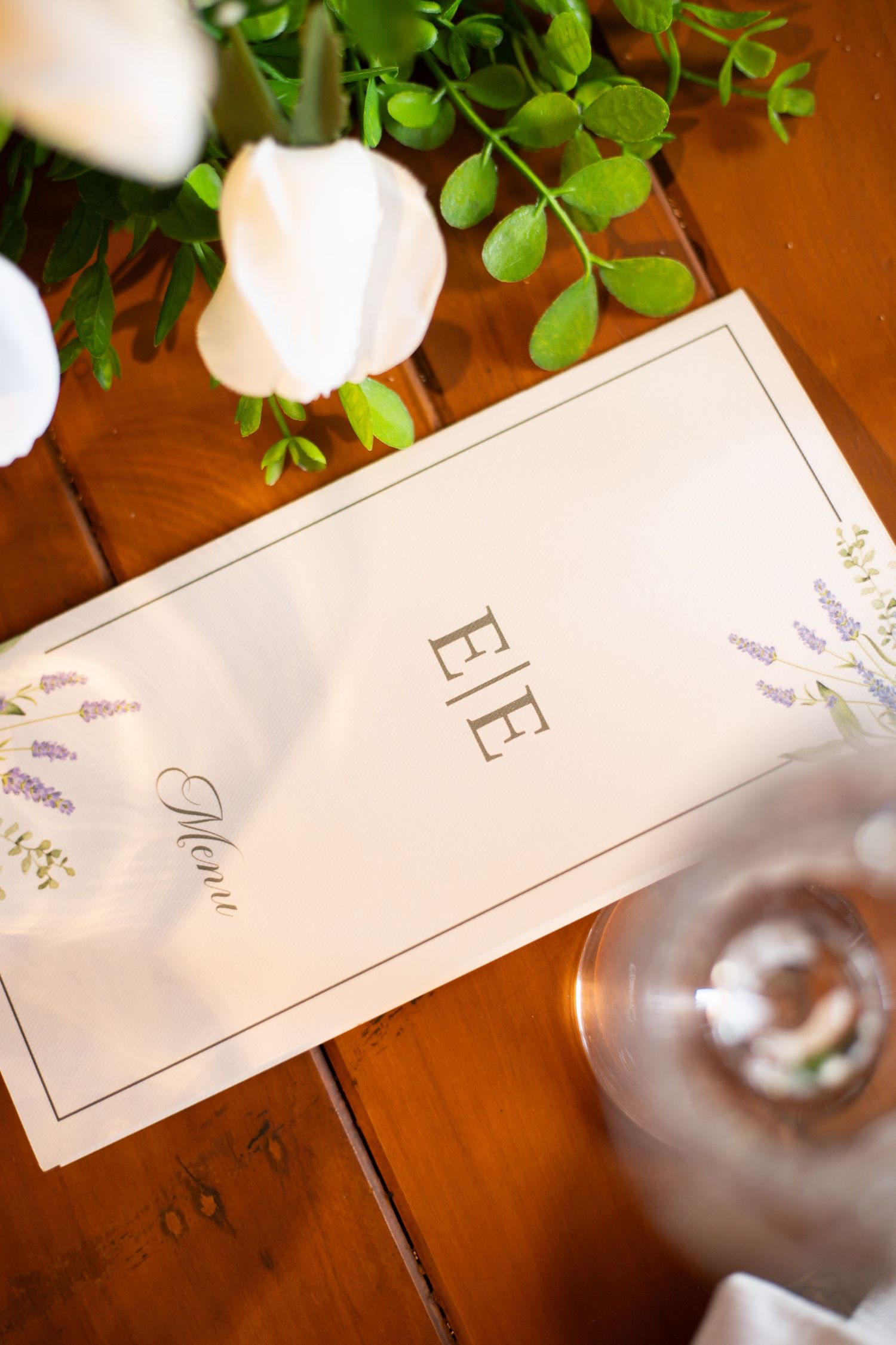Elegant wedding menu card with monogram EIE on wooden table, surrounded by white roses and green foliage.