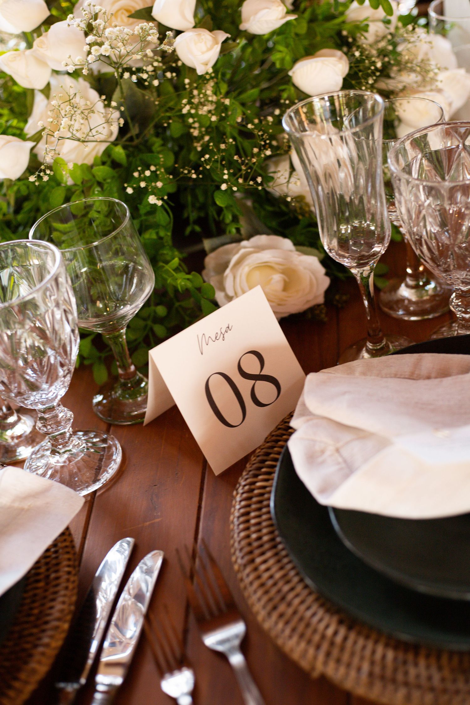Elegant wedding table setting with white roses, crystal glasses, wicker chargers, and a table number card reading 08.