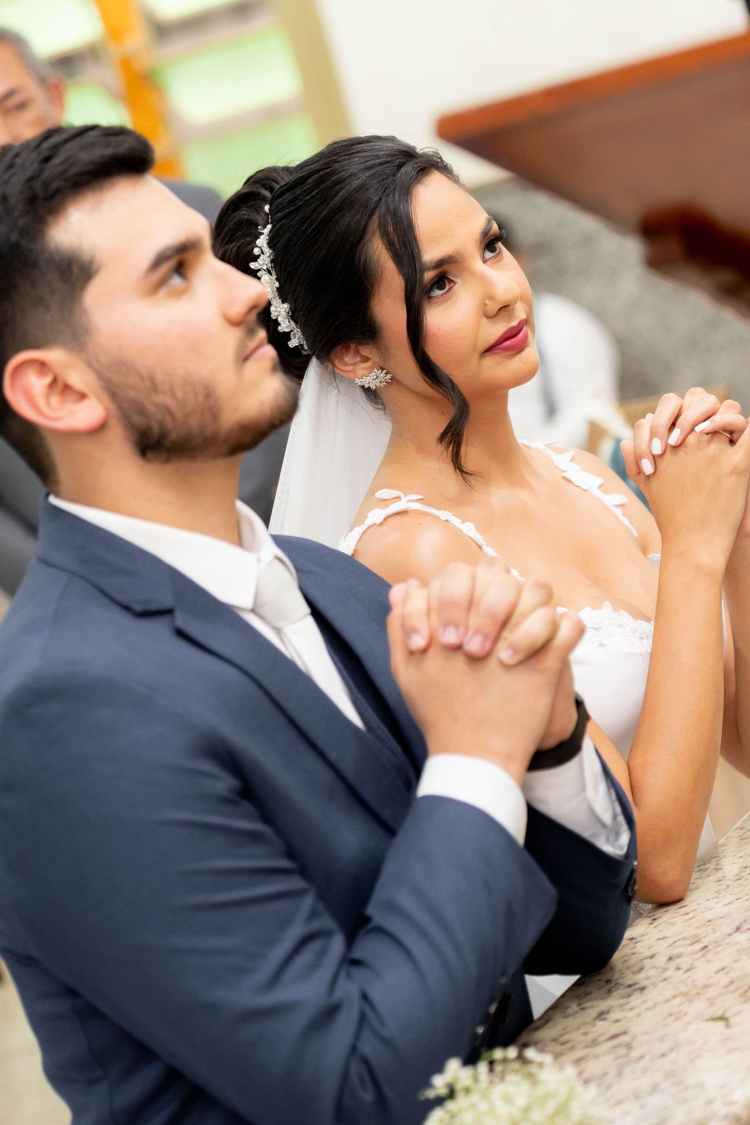 A bride and groom pray together at the altar during their wedding ceremony, dressed in a white gown and blue suit.