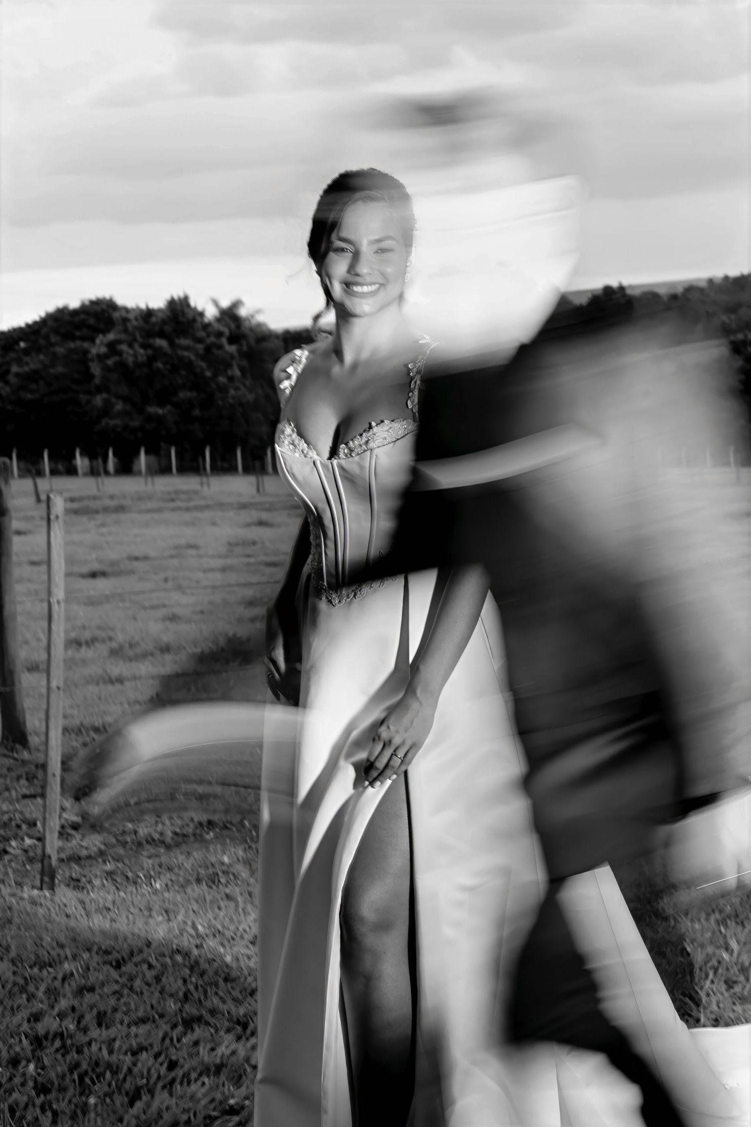 Black and white photo of a smiling bride in a gown outdoors, with a motion-blurred figure passing by.