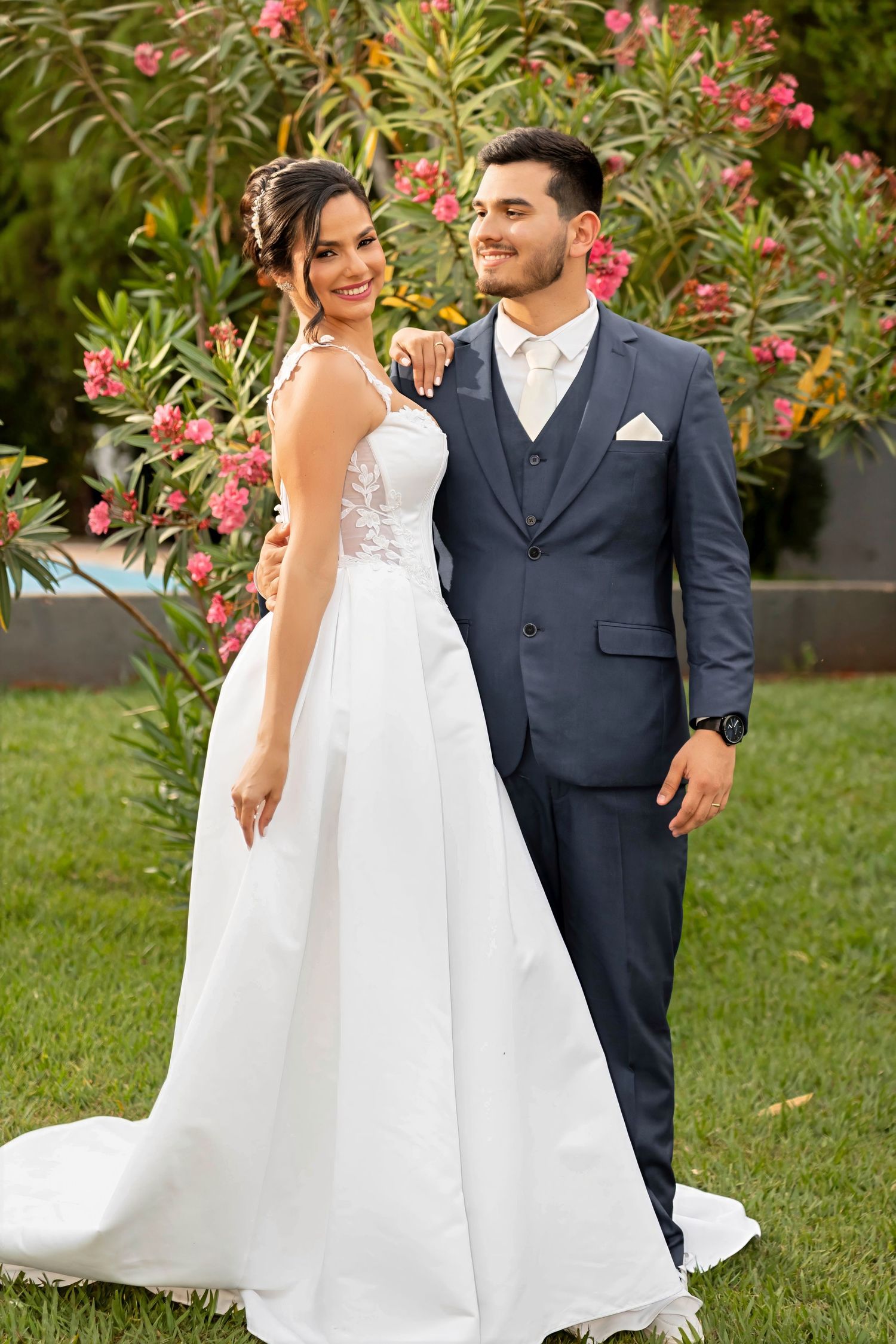Bride in white gown and groom in navy suit pose together outdoors among pink flowering bushes on their wedding day.