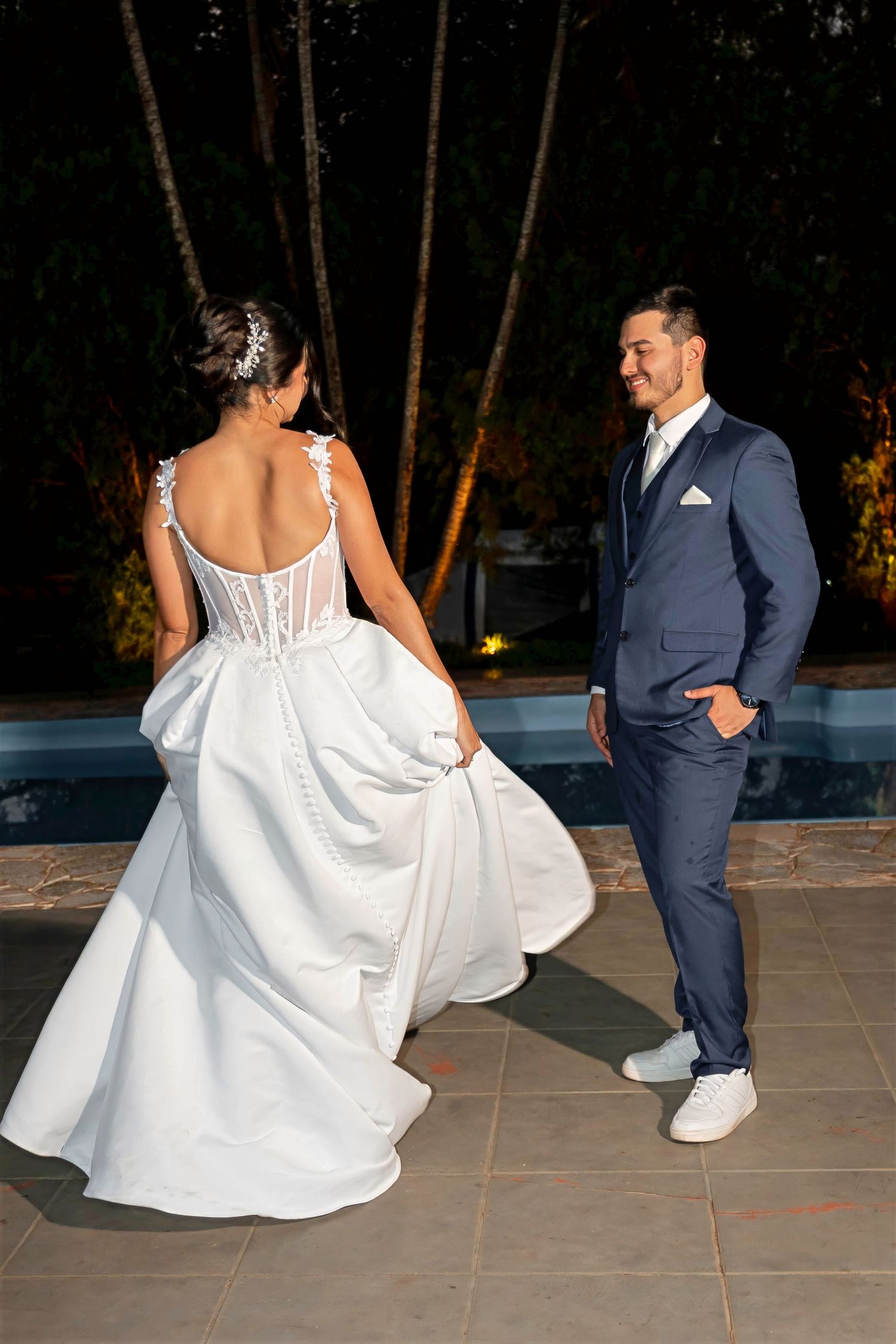 Bride in white gown twirls near pool as groom in navy suit smiles at her during outdoor evening wedding.