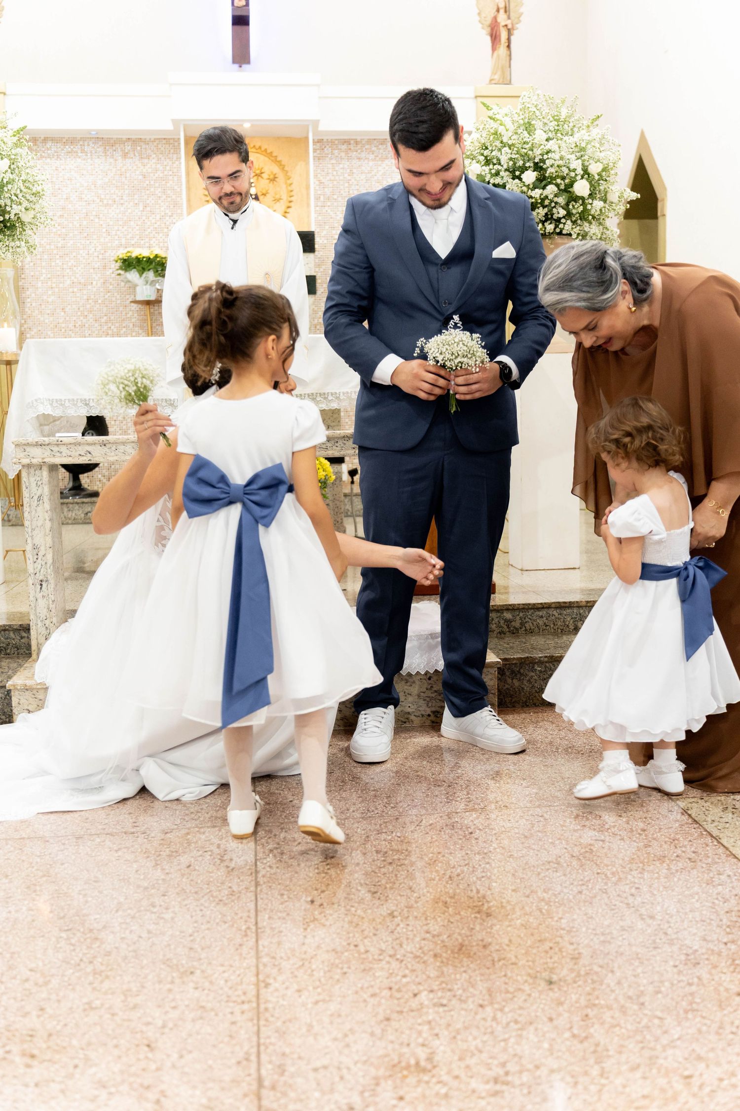 Groom in navy suit interacts with flower girls in white dresses with blue bows during a church wedding ceremony.