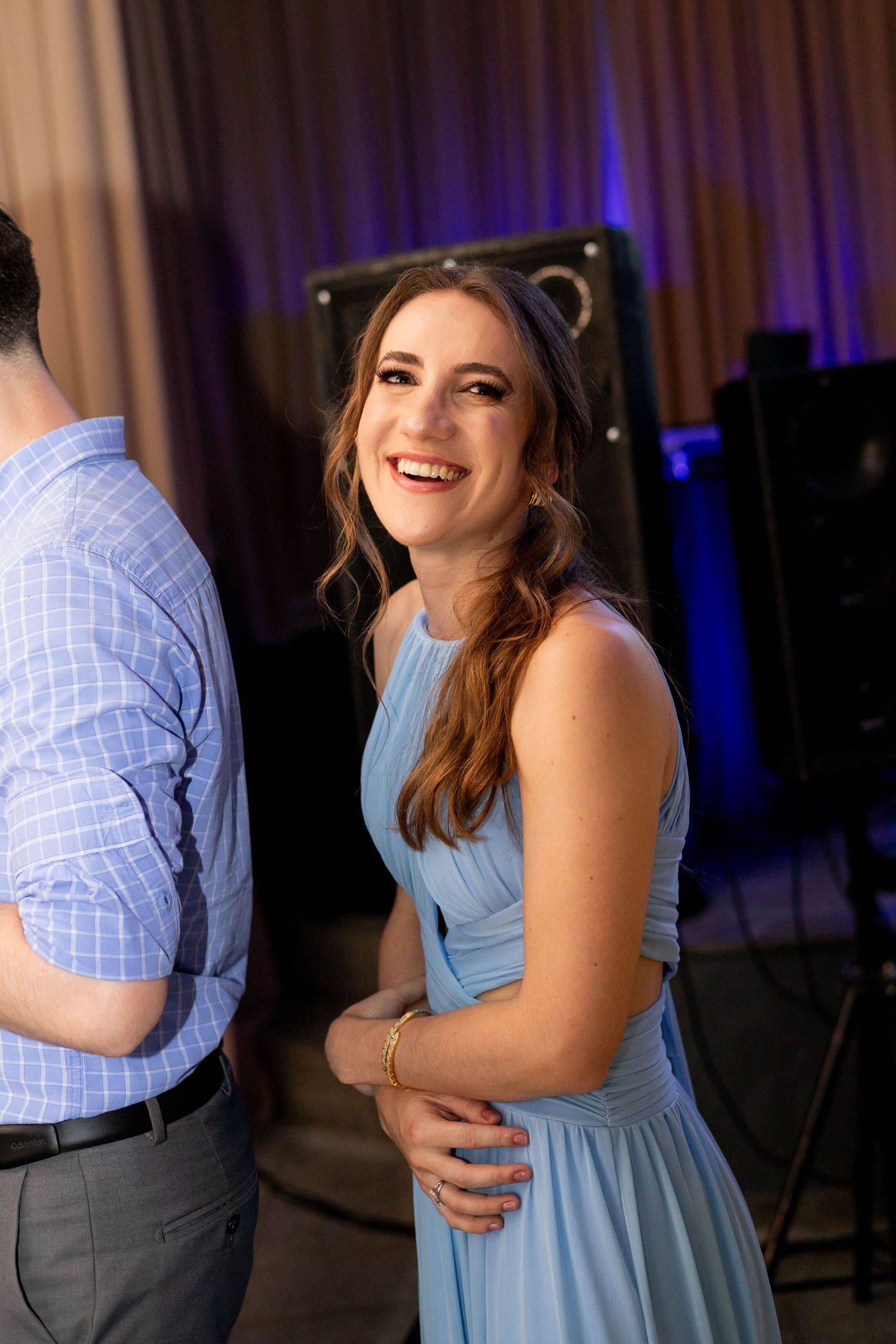 A smiling woman in a light blue dress enjoys a celebration event, with purple lighting and speakers in the background.