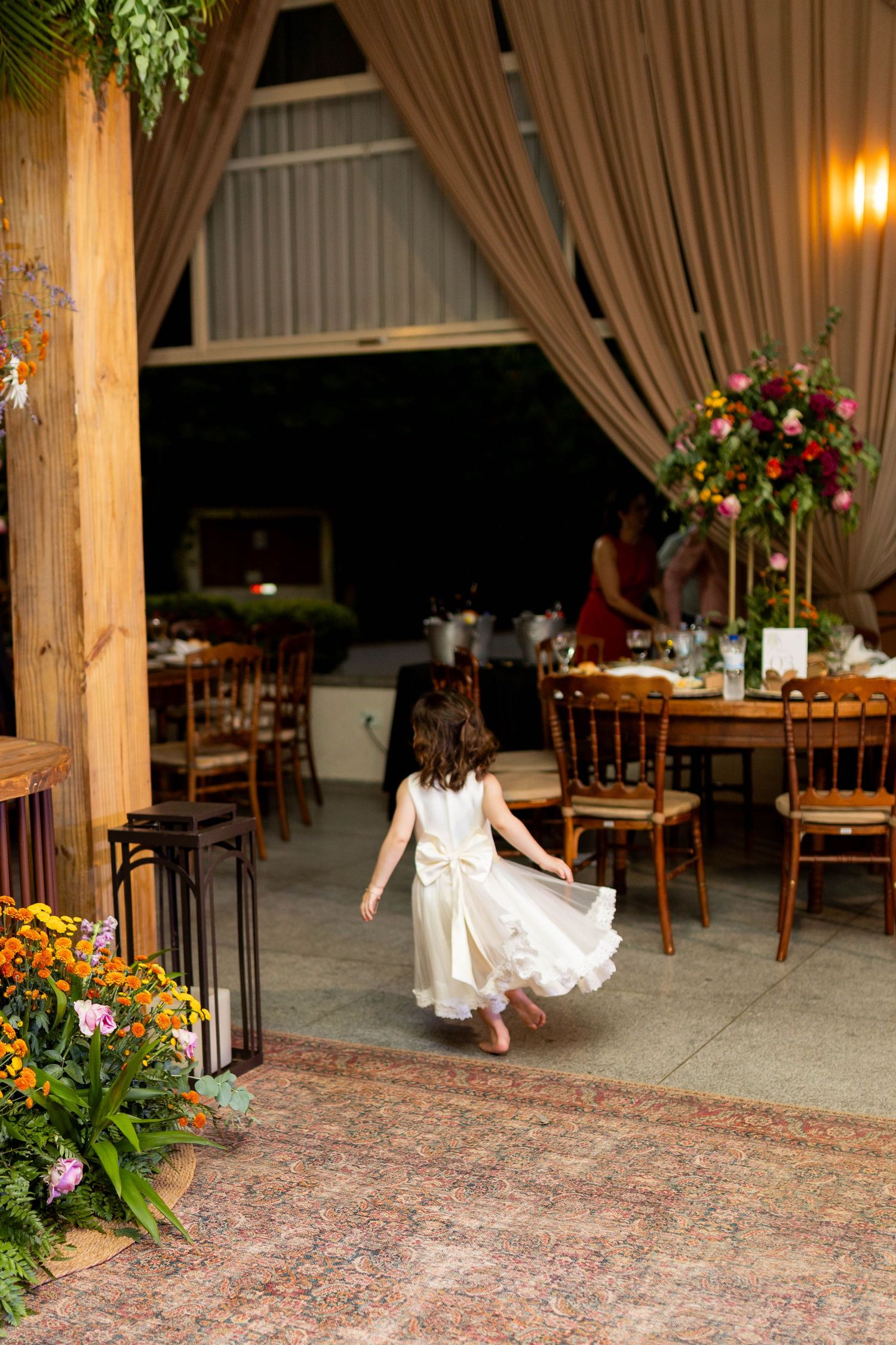 A flower girl in a white dress twirls at a wedding reception venue decorated with floral arrangements and draped curtains.