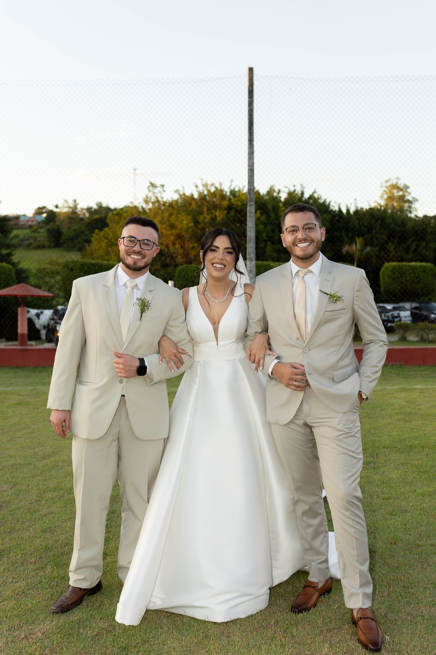 Bride in white gown poses with two groomsmen in beige suits at outdoor wedding venue.